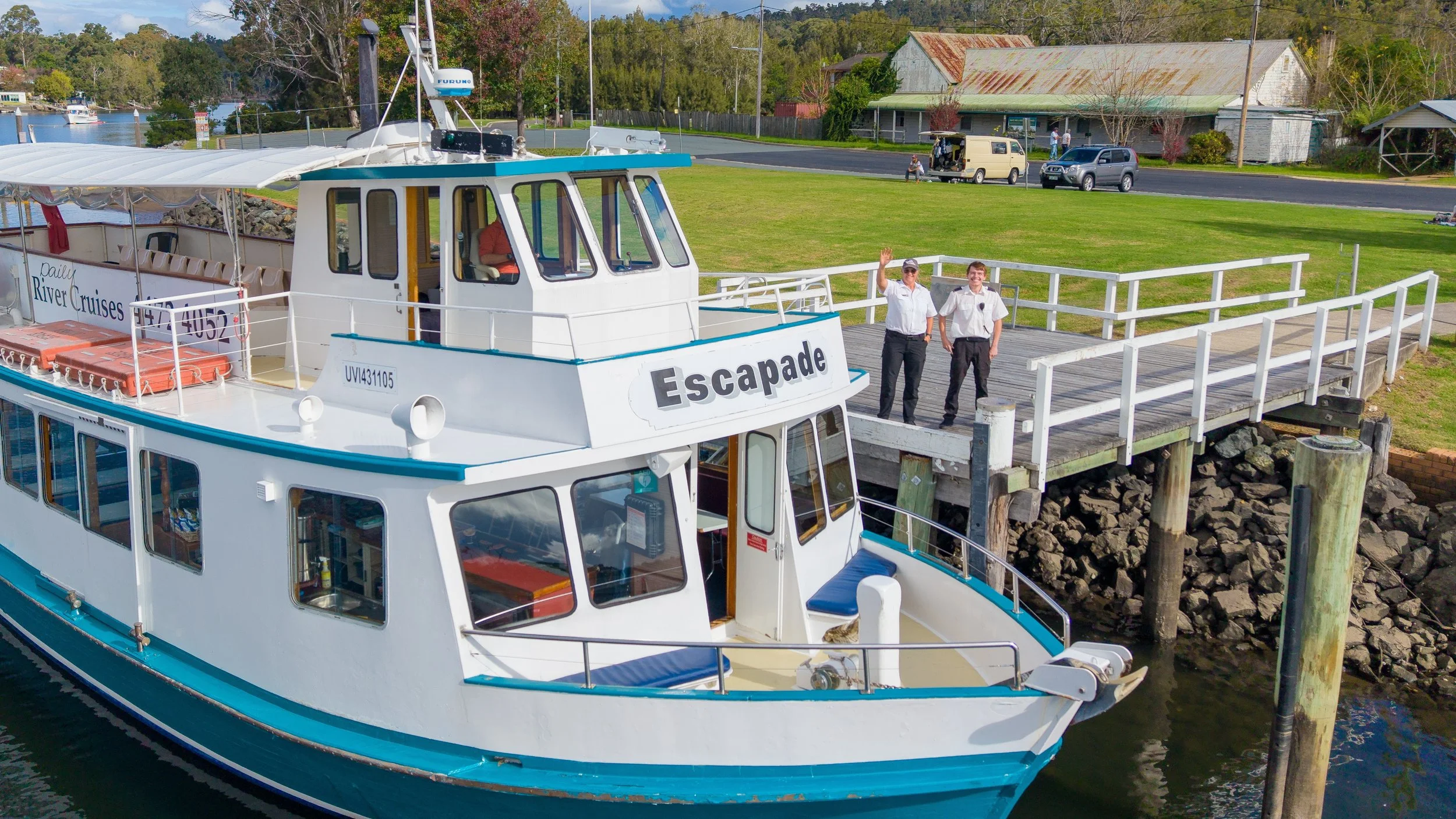 A white boat named Escapade docked at a pier with two crew members waving. In the background, there is a grassy area with a few trees, parked cars, and an old barn with a rusty roof.
