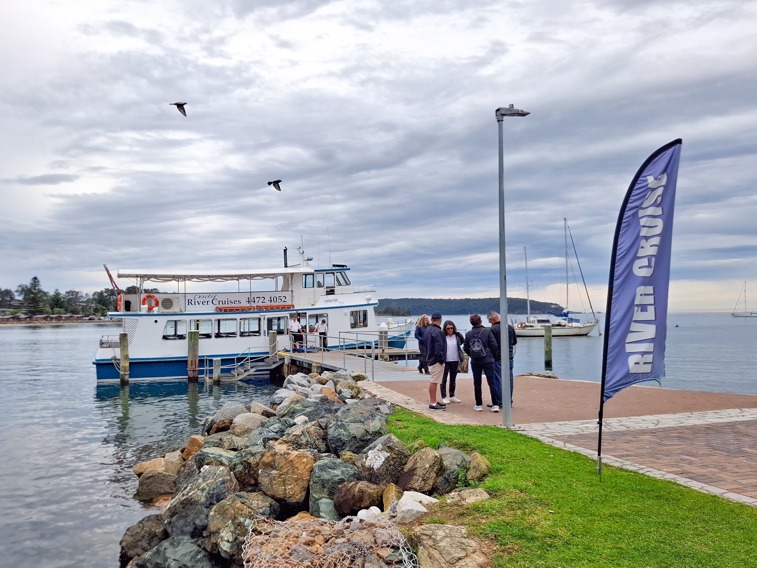 People standing near a docked boat named 'Raile River Cruises' at a marina with overcast skies, sailboats in the water, and a flag with partially visible text.