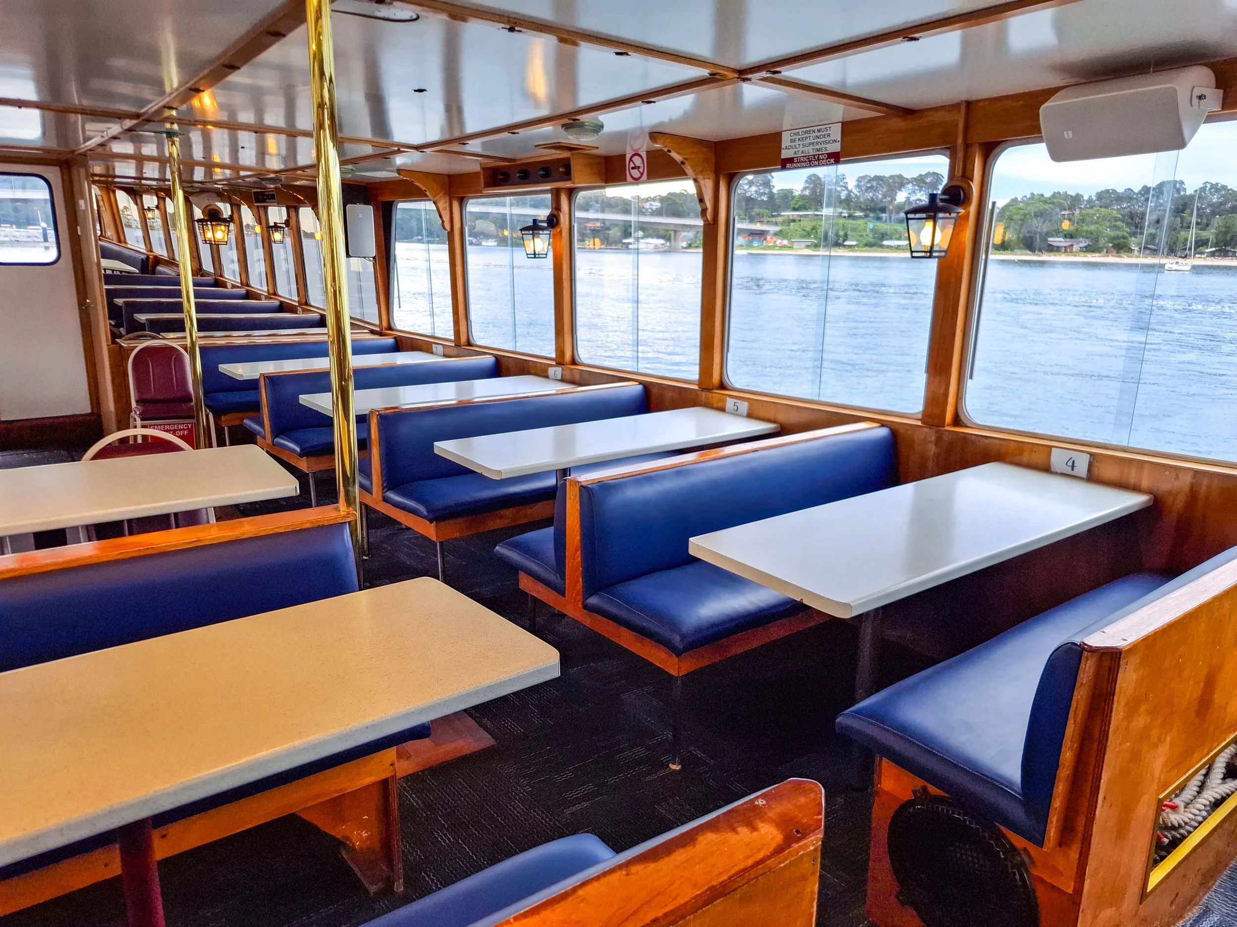 Interior of a boat dining area with blue cushioned seats and white tables, large windows showing water and greenery outside, and warm wooden finishes.