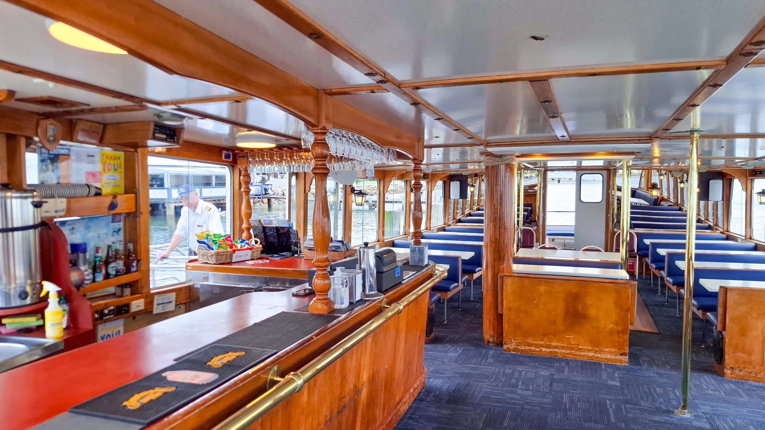 Interior of a boat's dining area with blue seats, wooden accents, and large windows showing water outside. A bar area with glasses, snacks, and a person in a uniform is visible.