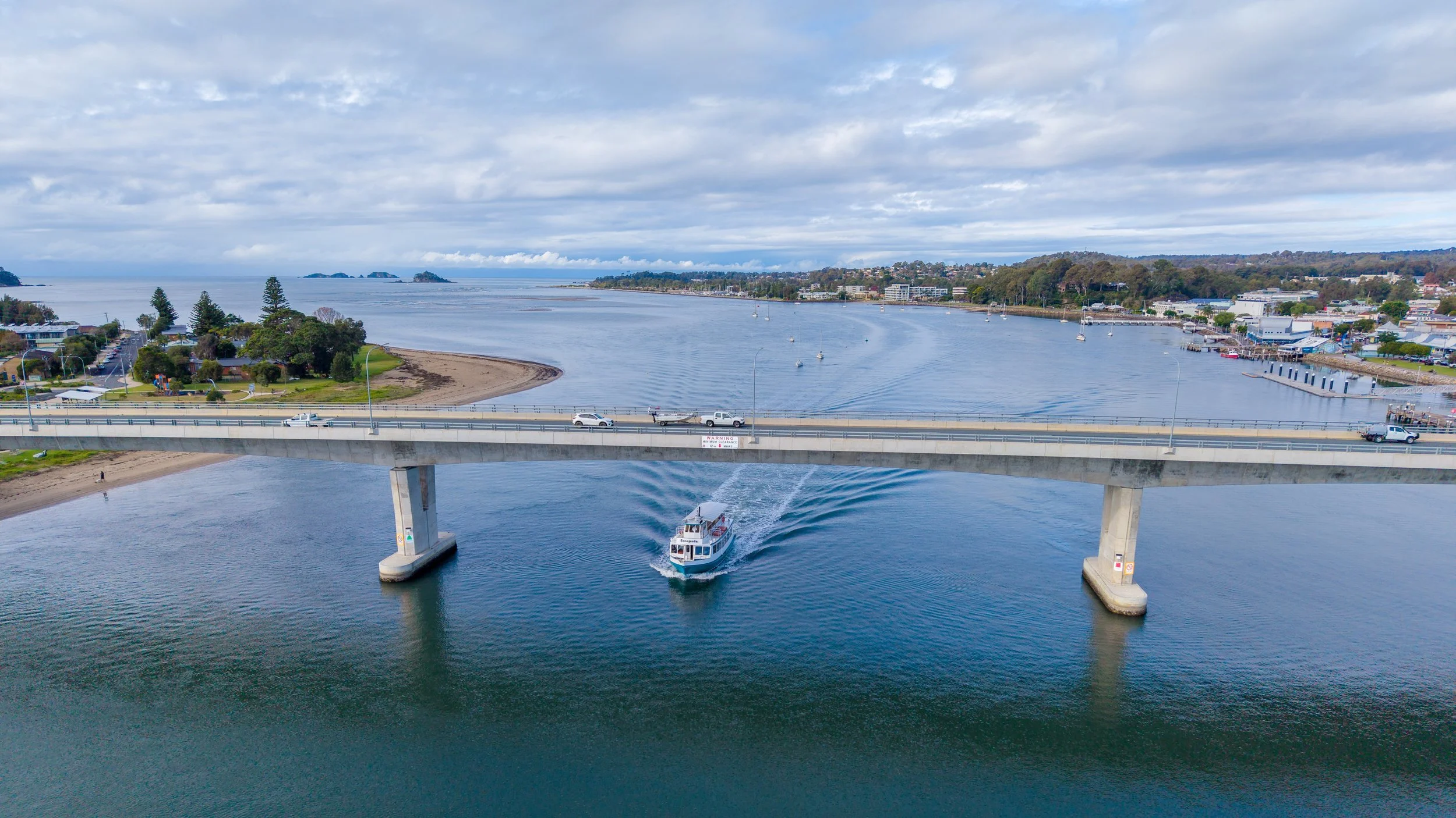 Aerial view of a bay with a bridge crossing over the water, a boat sailing underneath, and a city with buildings along the shoreline in the background.
