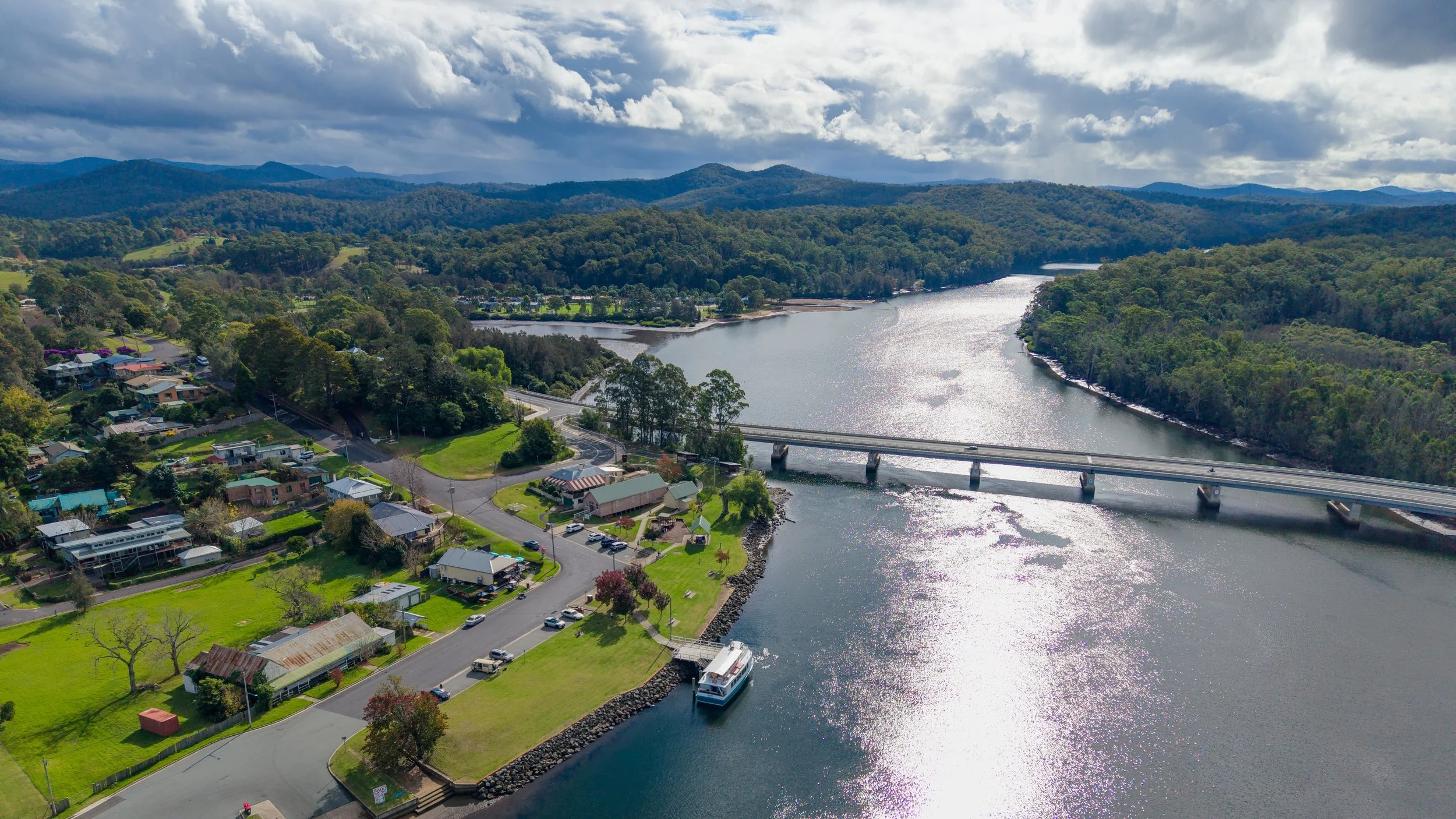 Aerial view of a river flowing through a town surrounded by forested hills and mountains, with a bridge crossing over the river and houses along the shoreline under partly cloudy skies.