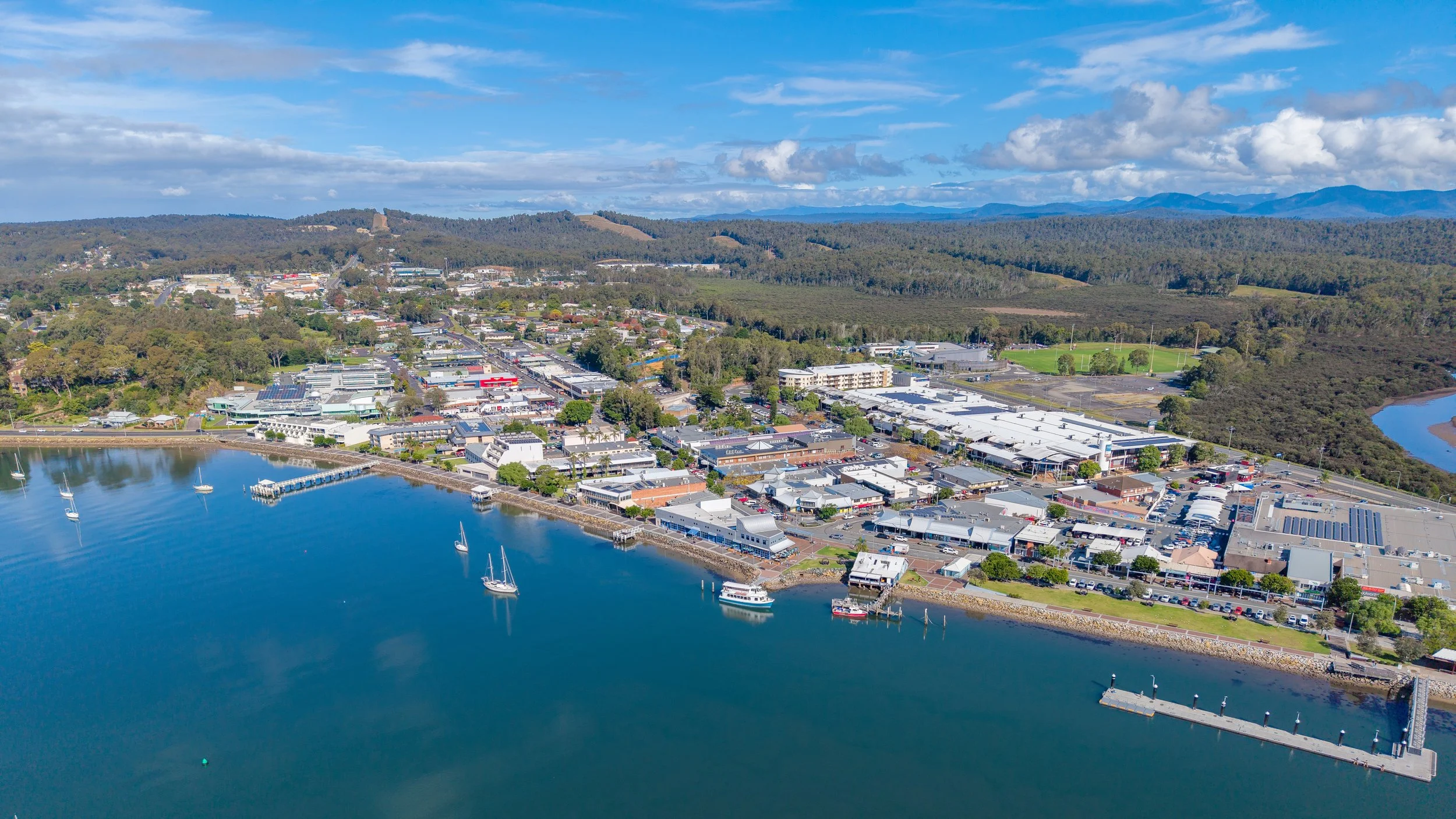 Aerial view of a lakeside town with boats docked at a marina, surrounded by commercial and residential buildings, green hills, and distant mountains under a partly cloudy sky.