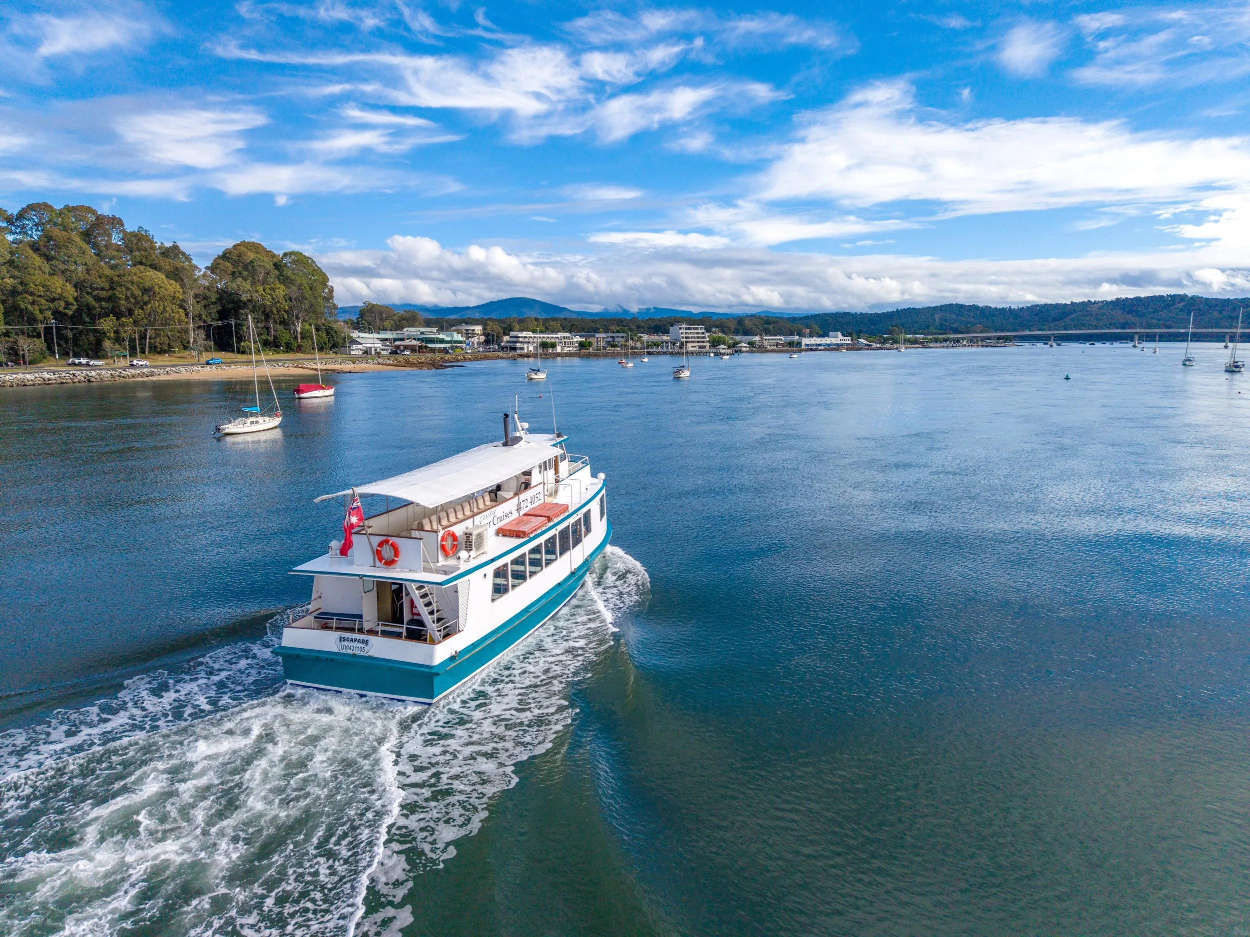 A white boat named 'Escapade' sailing on calm water with multiple sailboats anchored nearby and a shoreline lined with buildings and trees in the background, under a partly cloudy blue sky.