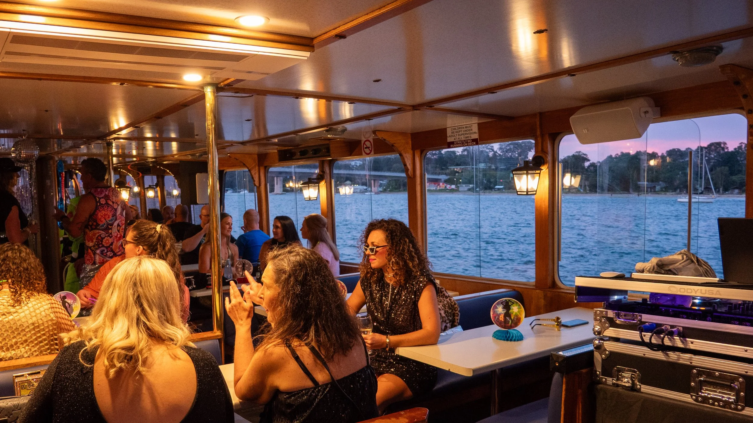 Interior of a boat with passengers seated, enjoying a social gathering during sunset, with water and trees visible through the windows.
