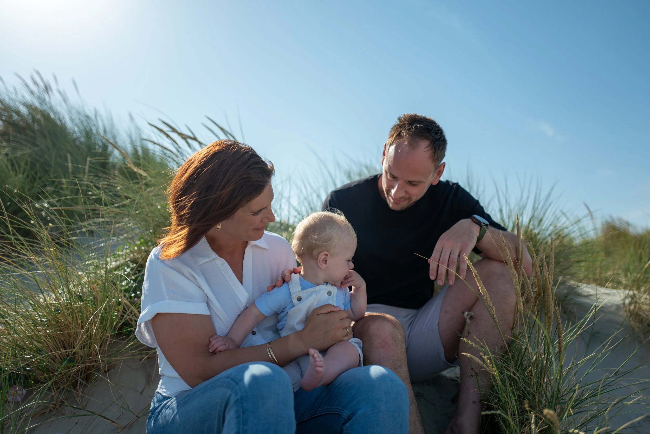 A family of four, including a woman, a man, and two young children, sitting on the beach among tall grass under a clear blue sky, enjoying a sunny day.