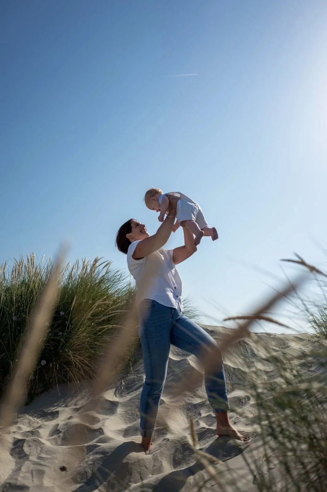 A woman lifting a baby in the air on a sandy beach with tall grass, under a clear blue sky.