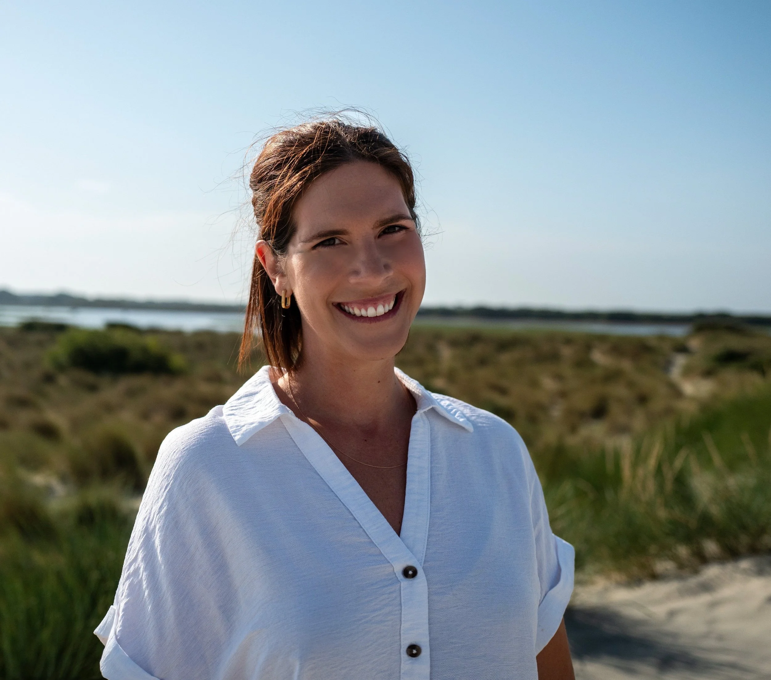 Close-up of a woman smiling outdoors near water, wearing a white shirt and gold hoop earrings.