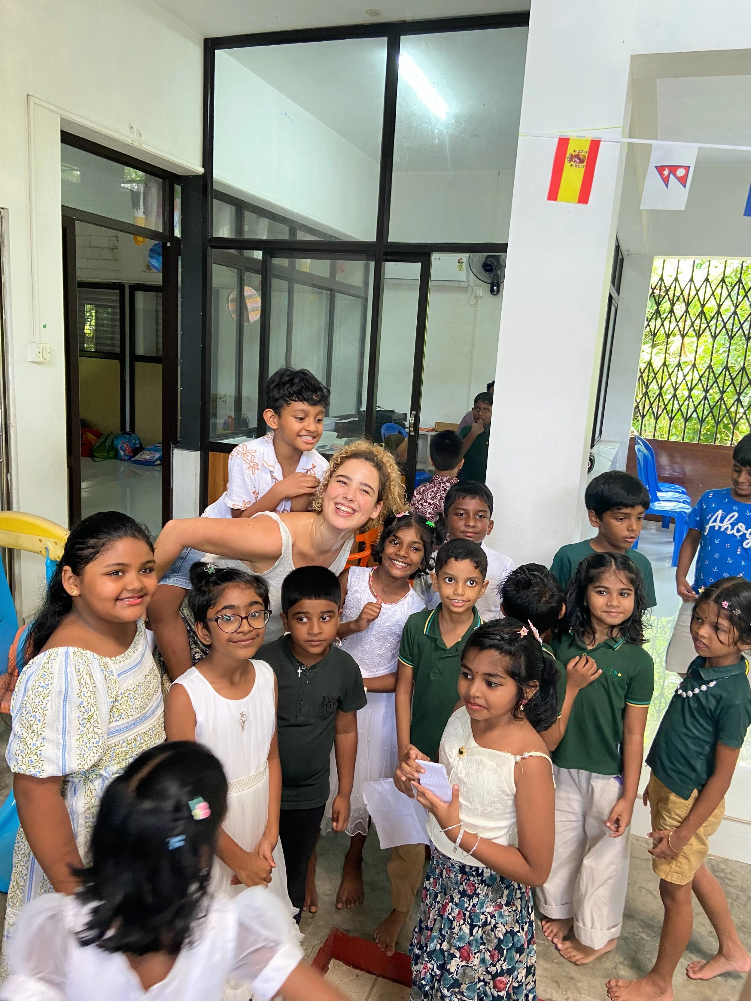 A group of children and a young woman are gathered inside a room, smiling and posing for the camera. Some children are wearing casual clothing, and the environment suggests a celebratory or social event.
