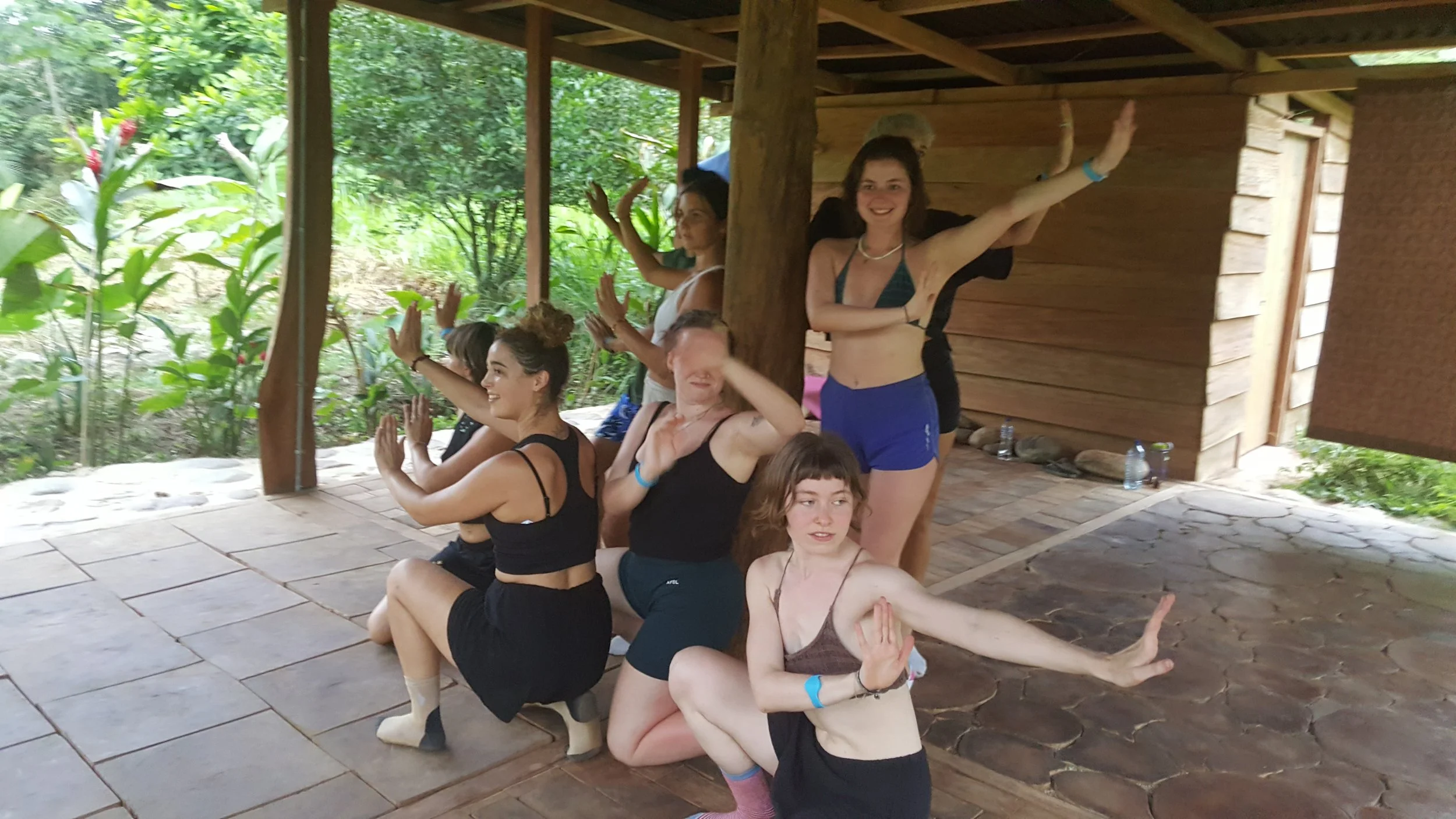 Group of young women in a dance class in a staggered formation on a covered outdoor patio with a natural forest background.