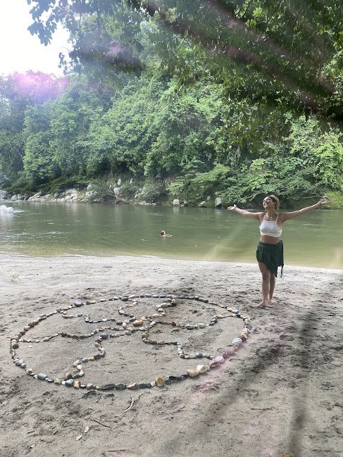Person standing on the beach near a river, arms outstretched, with a sun shape made of rocks on the sand. There are trees and another person swimming in the river in the background.