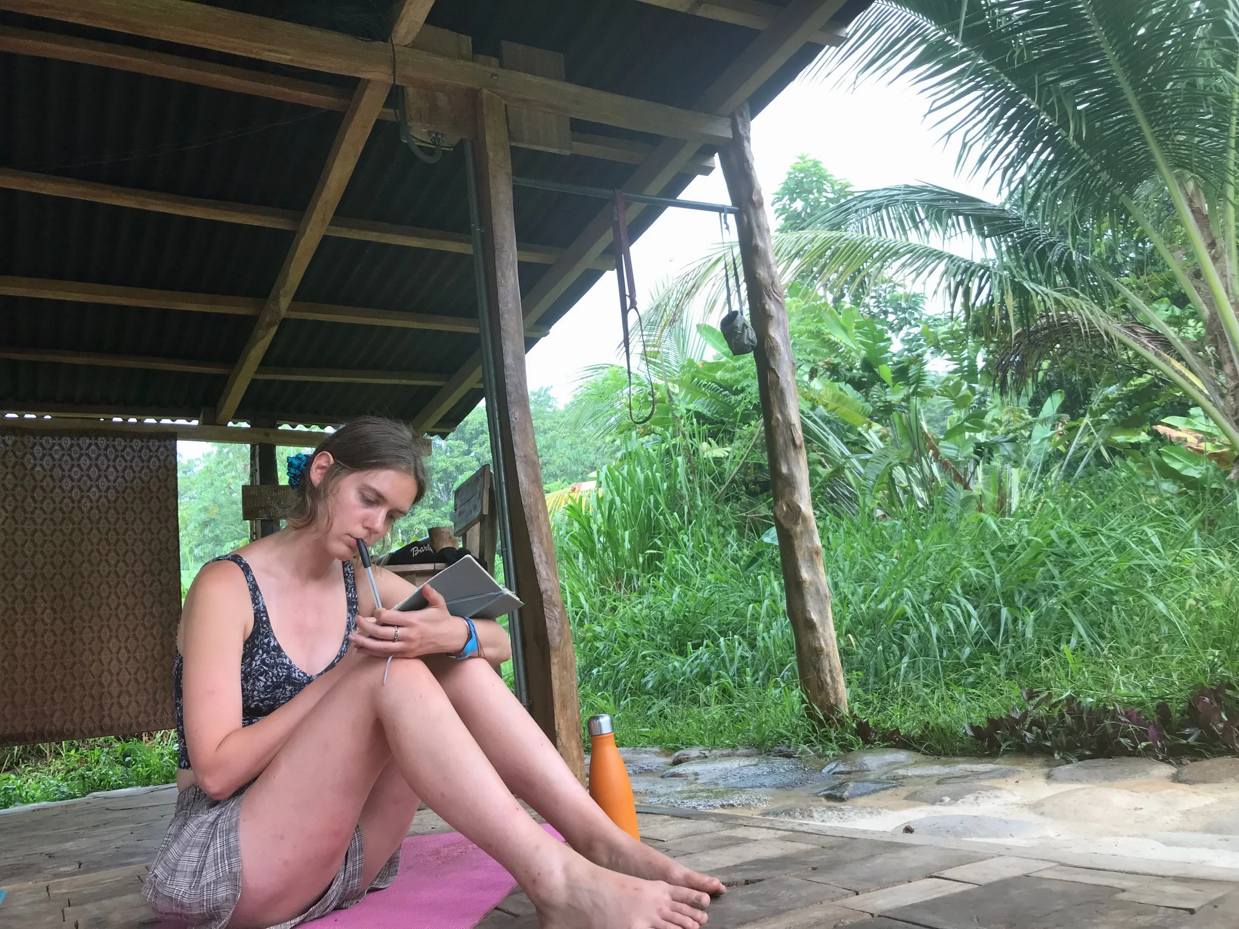 A woman writing surrounded by lush green tropical vegetation. She is sitting on a pink yoga mat, wearing a sleeveless top and shorts, focusing on her breathing or meditation with her eyes closed and holding her leg in a stretching pose.