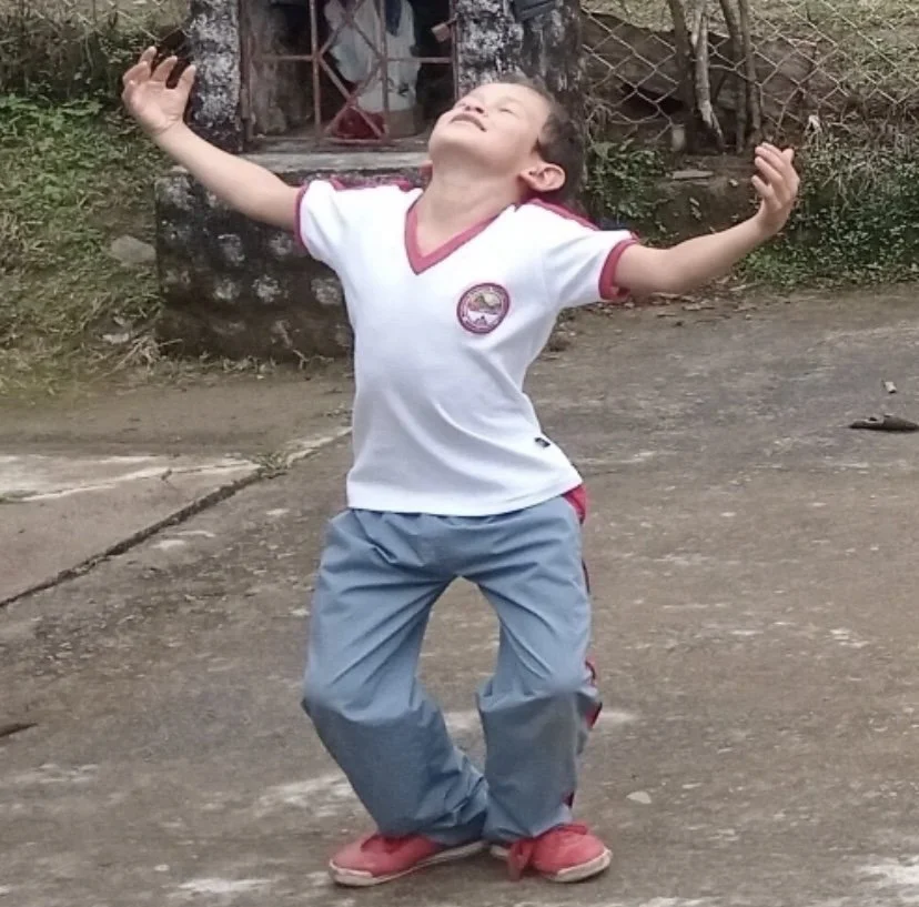 Child in a white T-shirt with red trim and gray pants standing on a concrete surface with arms raised and head tilted back.