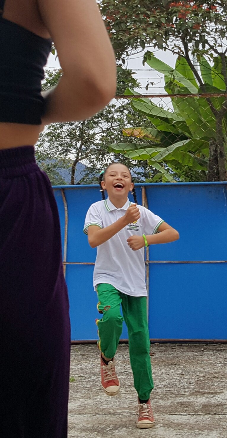 A young girl with pigtails smiling and laughing while running outdoors, wearing a white polo shirt and green pants, with a person partially visible in the foreground.