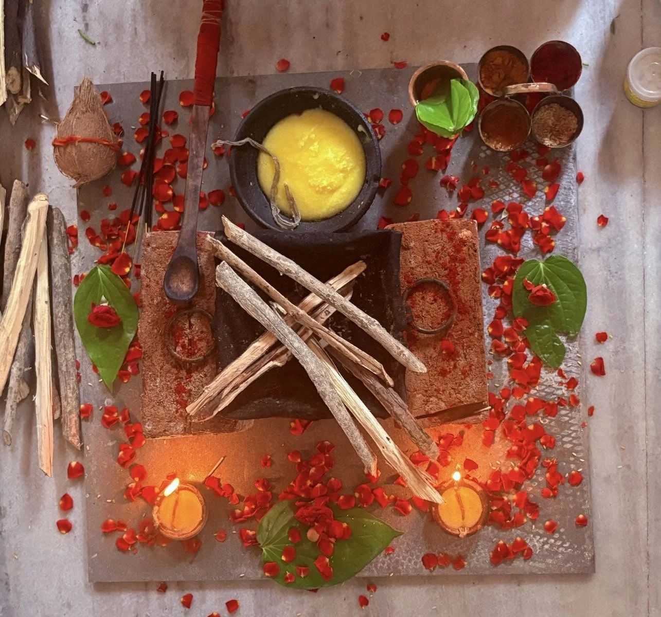 A small altar decorated with red flower petals, green leaves, and candles, featuring a clay fire pit with wooden sticks and a black pot with yellow soap or butter, surrounded by small containers and natural elements.
