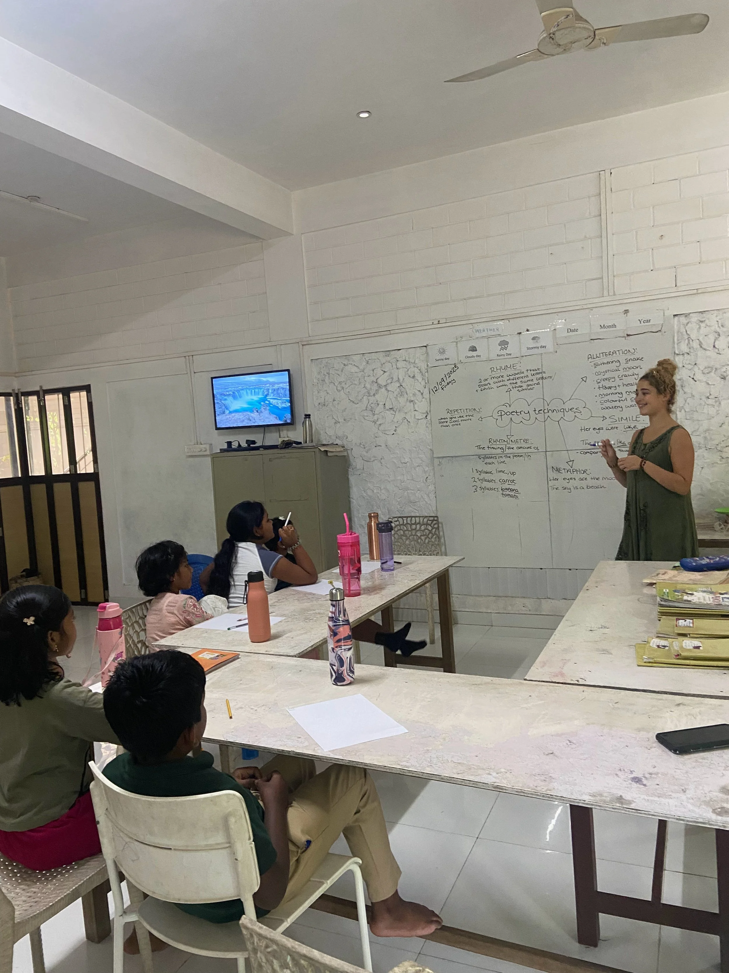 A classroom scene with a woman teaching students seated at tables, white brick wall with written notes and a monitor on the wall, ceiling fan, and white ceiling.