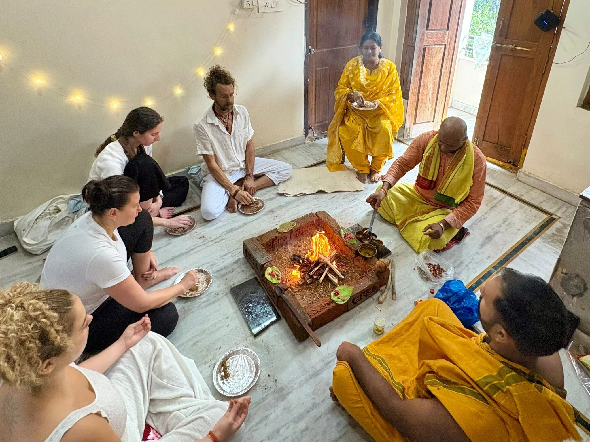People sitting cross-legged on the floor near a fire, engaging in a spiritual  ceremony. One person standing is dressed in a yellow traditional outfit, offering something to the fire.