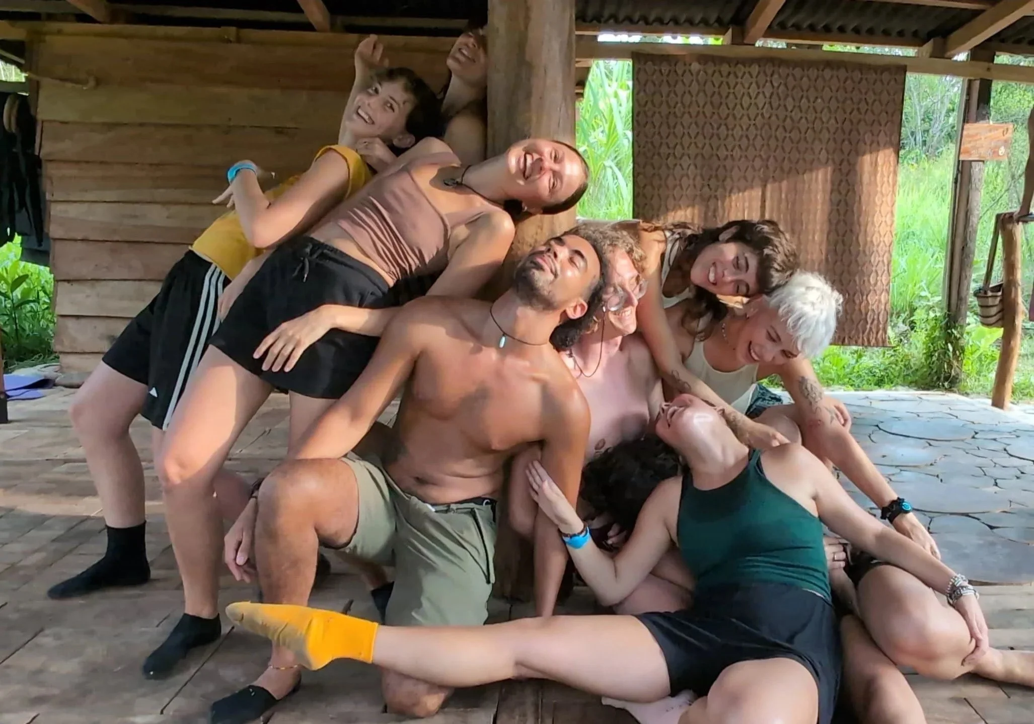 Group of happy people lying on a wooden floor, leaning against each other, in a large circle, in an outdoor setting surrounded by greenery, with some individuals wearing wristbands and casual clothing.
