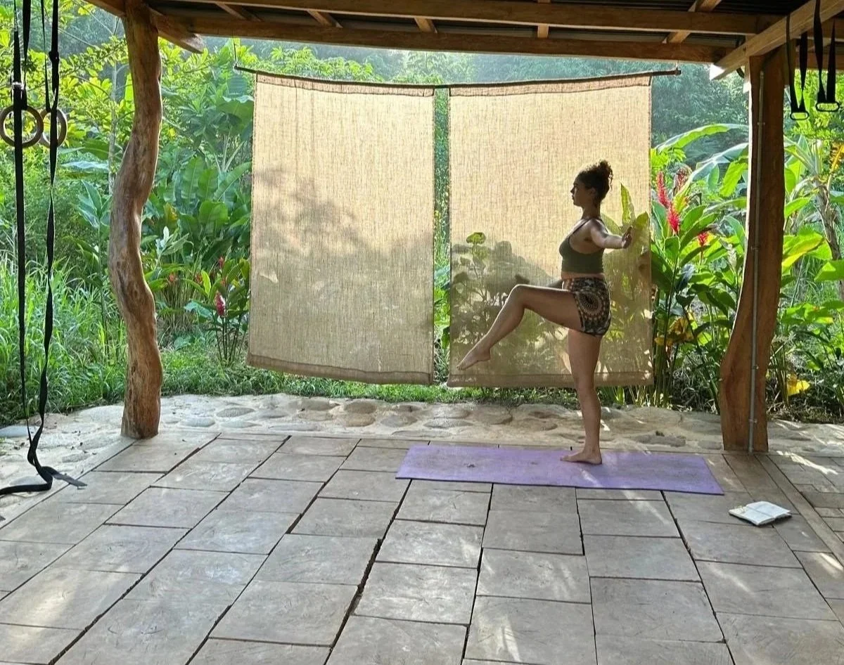 A woman practicing yoga outdoors on a purple mat, standing on one leg with her other leg raised and arms extended, on a wooden porch surrounded by lush green tropical plants.
