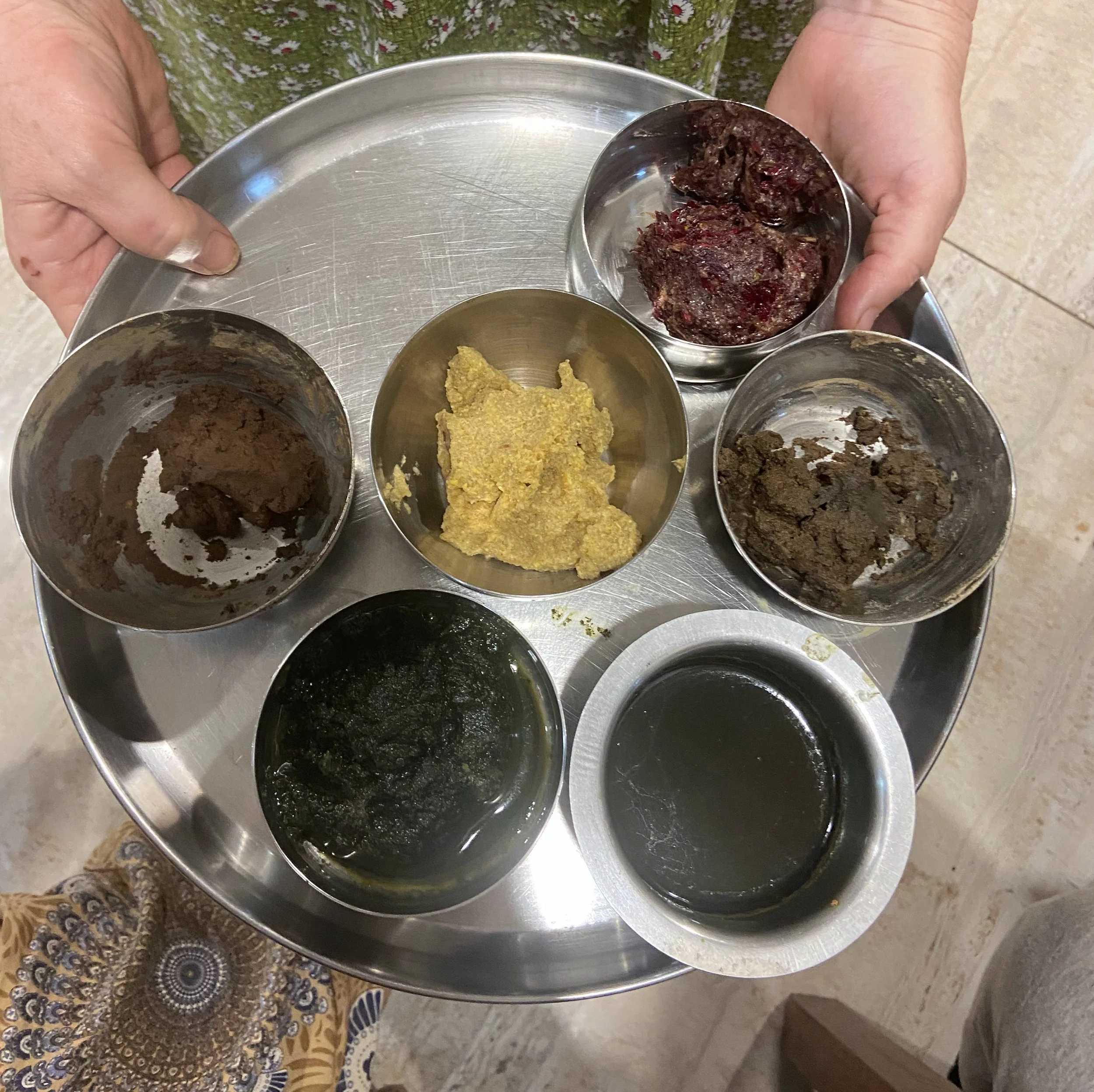 Person holding a metal tray with small cups containing different types of halva or sweet pastes, some dark and some yellow, on a speckled countertop.