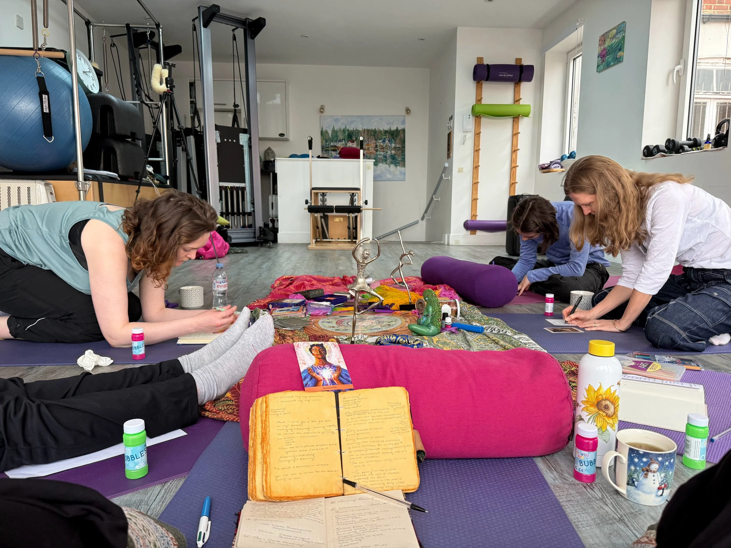 People sitting on the floor engaged in a group activity, surrounded by various art supplies, papers, and colorful cushions in a bright room with gym equipment and artwork on the walls.