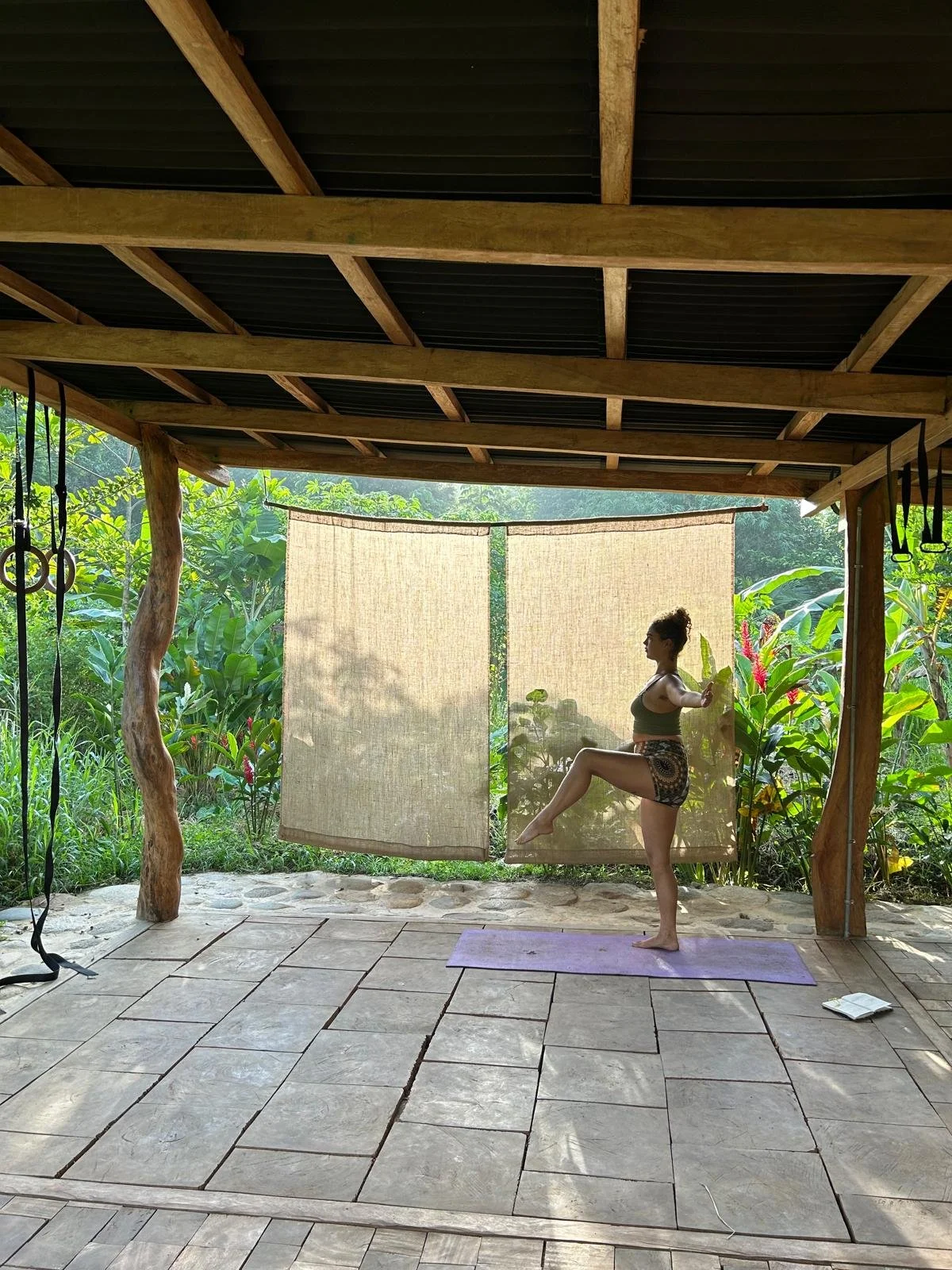 A woman practicing yoga outdoors on a purple mat in a covered, open-air structure surrounded by lush greenery and large tropical plants.
