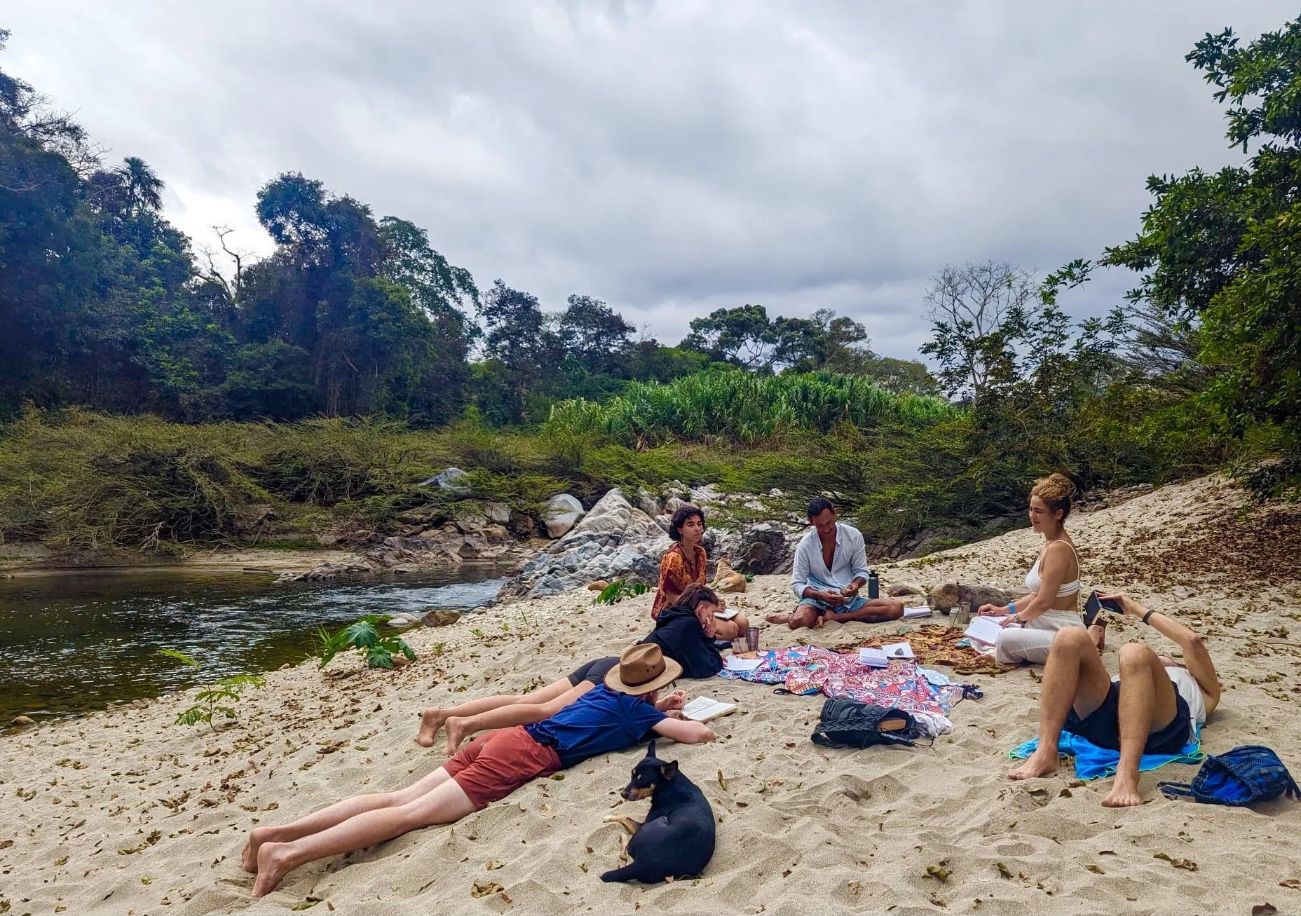 People sitting and lying on a sandy beach near a river, surrounded by green trees, with cloudy sky overhead.