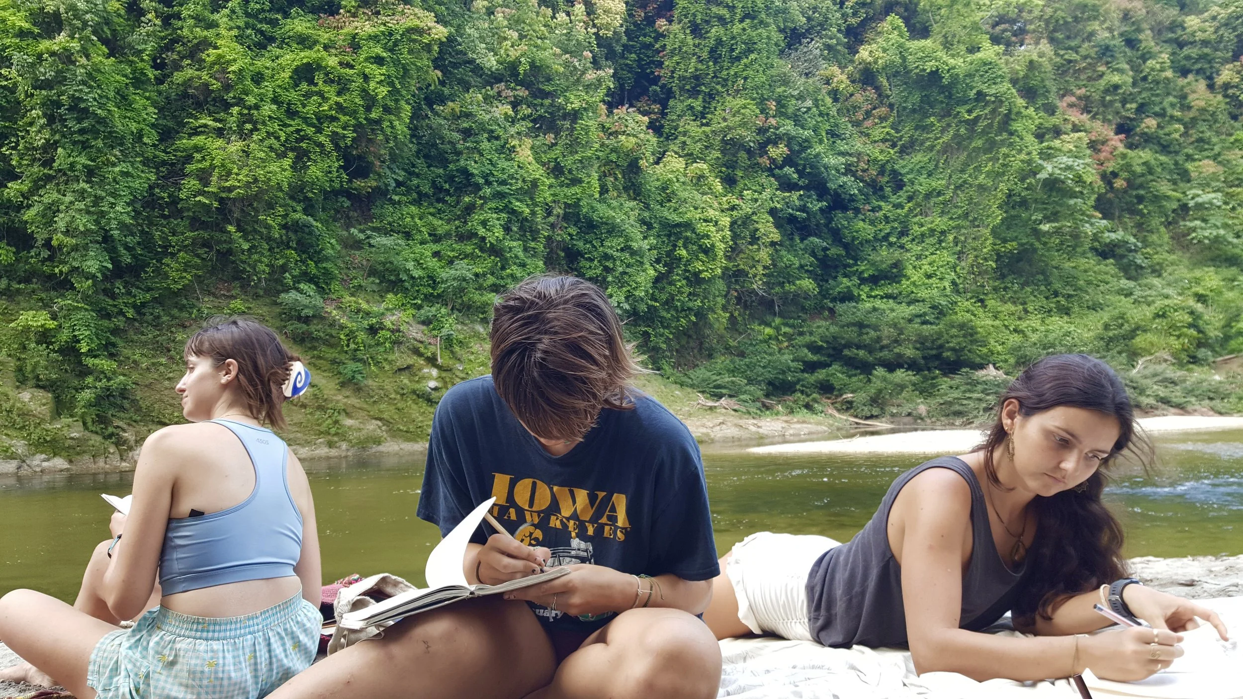 Three young women sitting on a riverbank near a forest, reading books and using their phones.