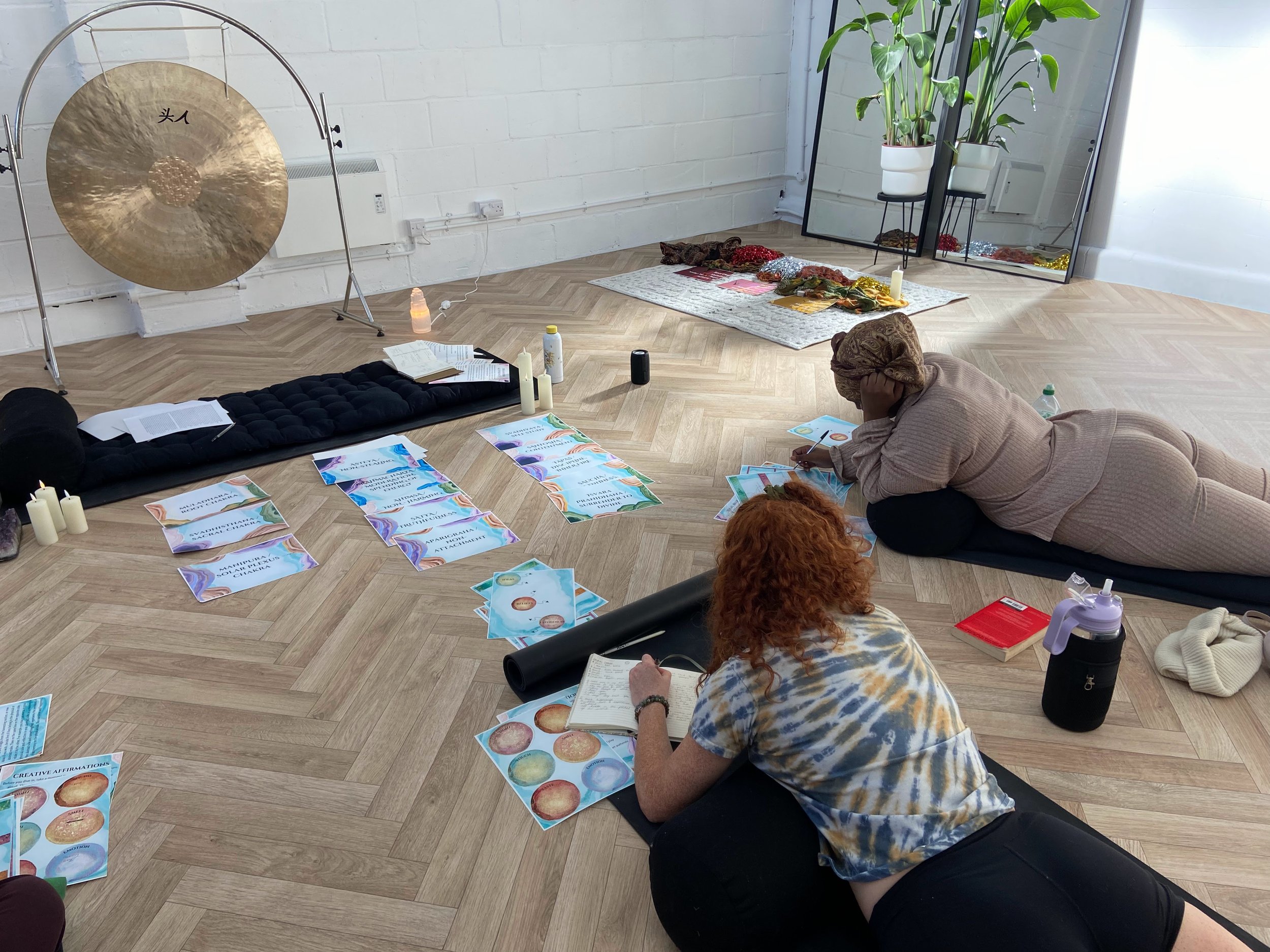 People participating in a workshop or class, lying on the floor with printed materials, candles, and a large gong, in a bright room with plants and a mirror.