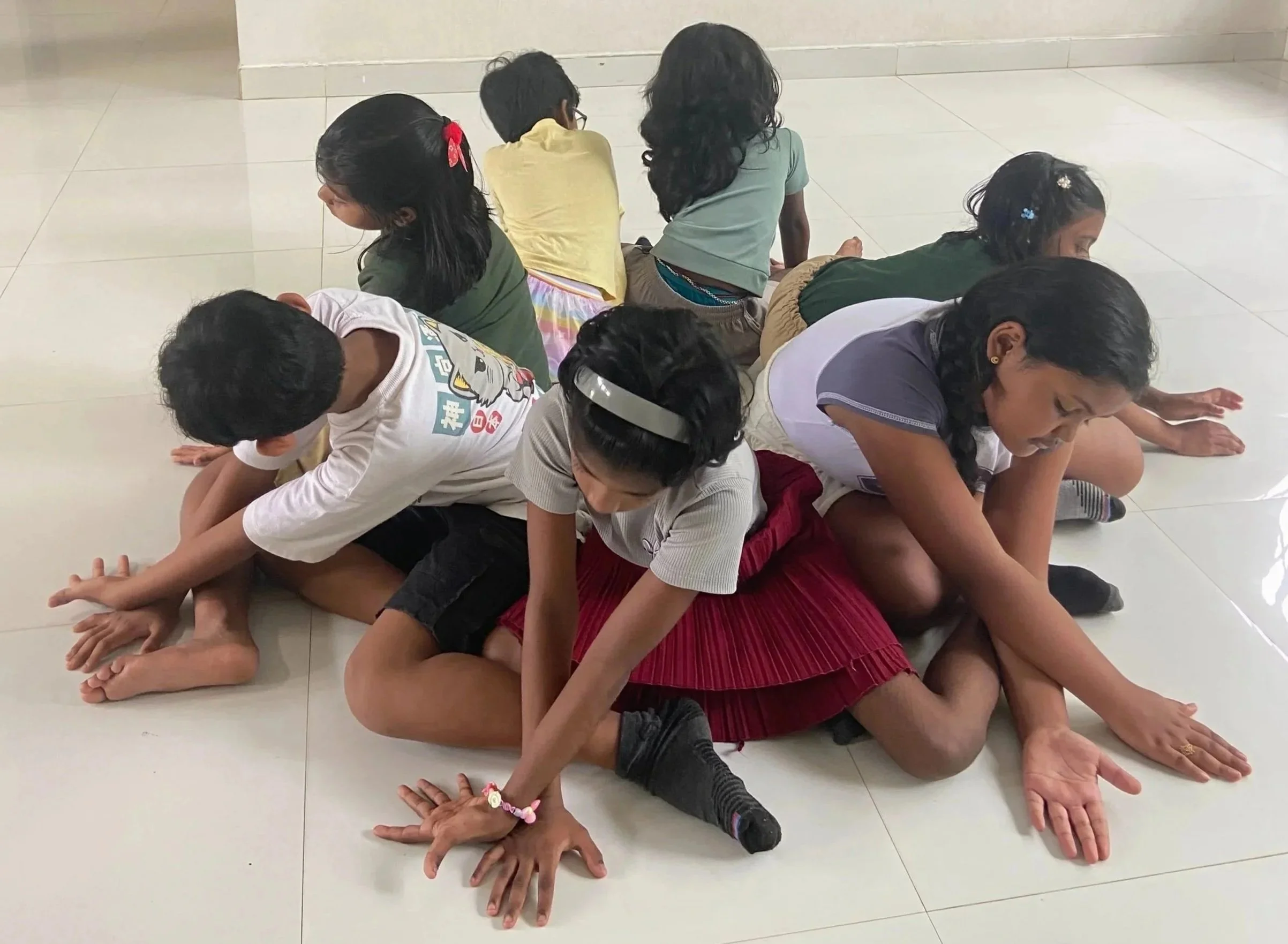 A group of children sitting on the floor, facing in different directions, with some leaning on each other, in a spacious room with tiled flooring and plain walls.
