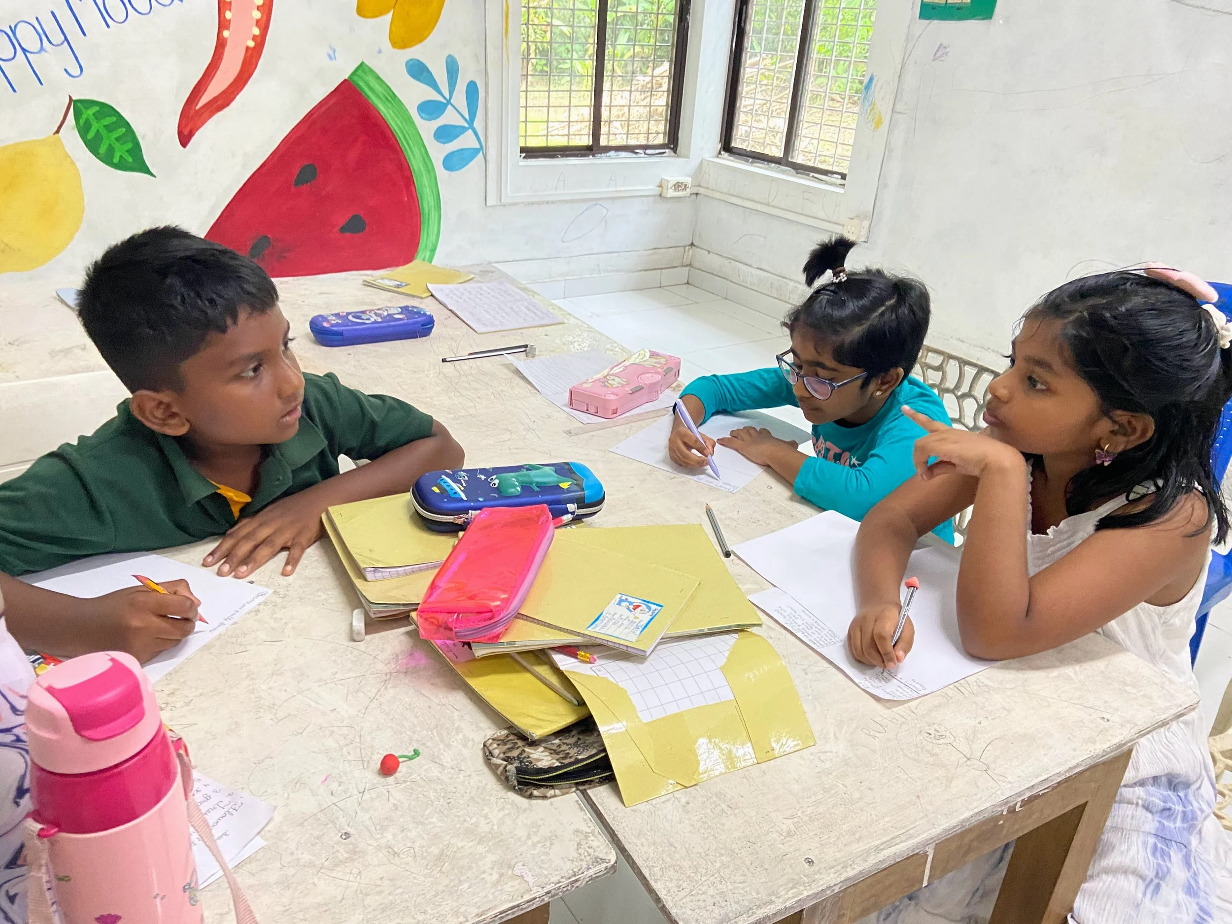 A group of children sitting around a table in a classroom, engaging in writing activities with notebooks and pens, with colorful artworks, including a large watermelon mural, on the walls and windows letting in natural light.