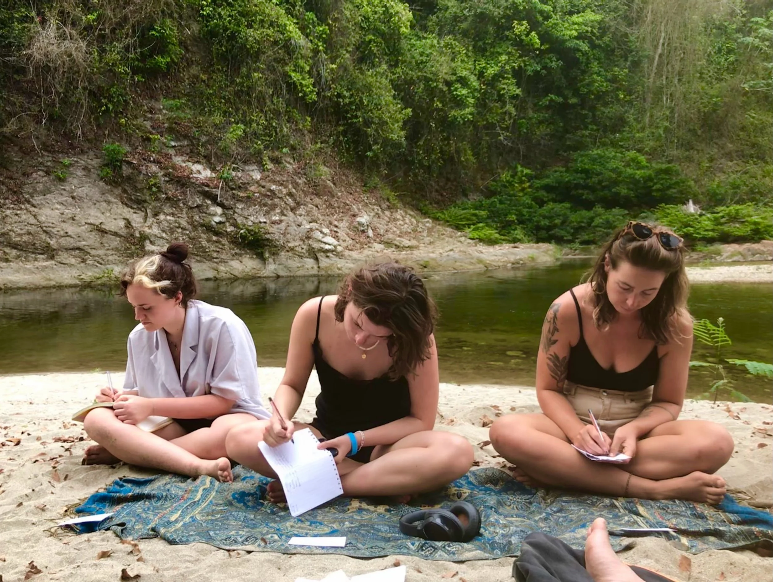 A group of three young women sitting cross-legged on a colorful blanket on the beach, writing  on notebooks during daytime, with a river and green trees in the background.