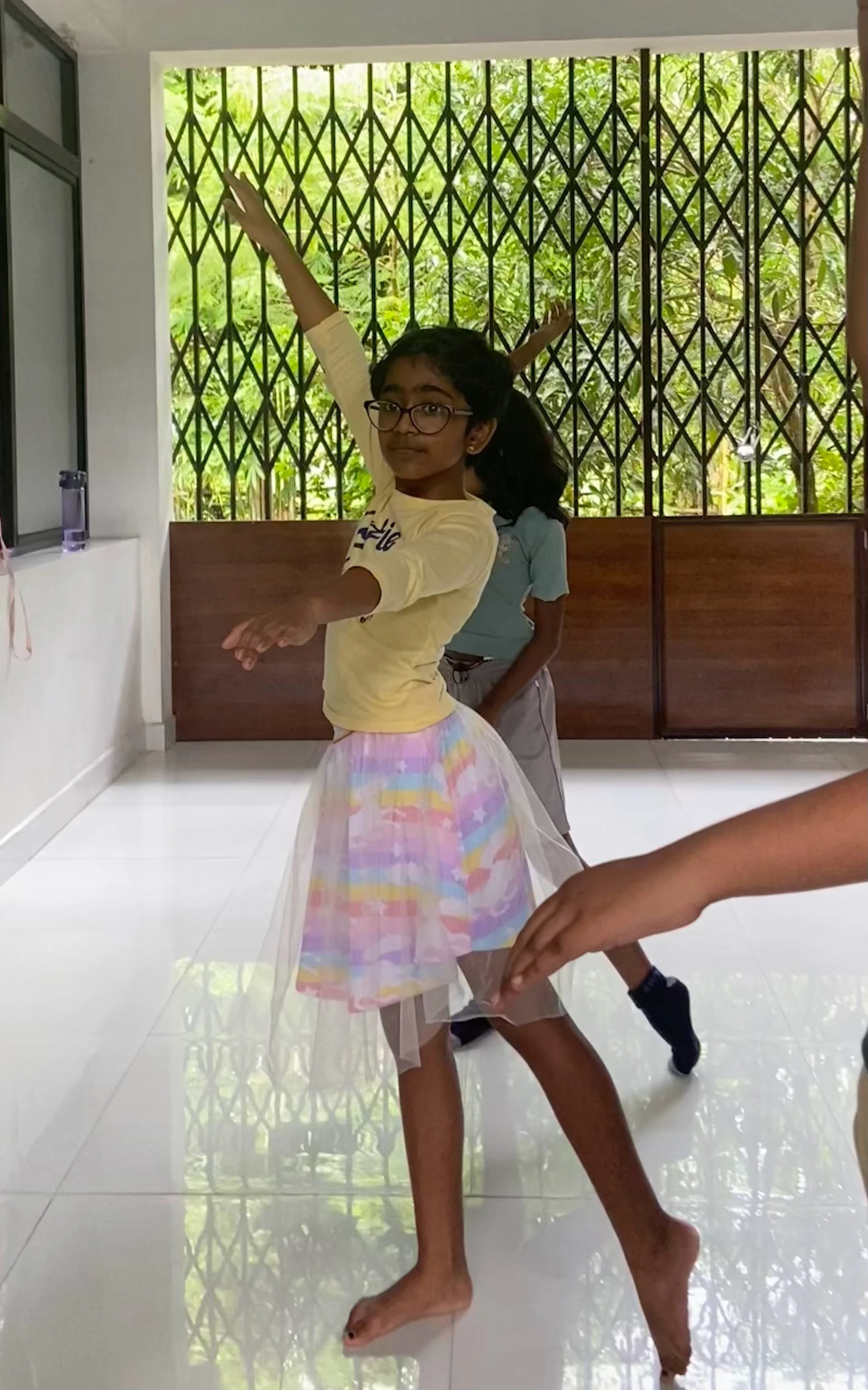 Children playing and dancing indoors near a window with metal bars, with green trees outside.