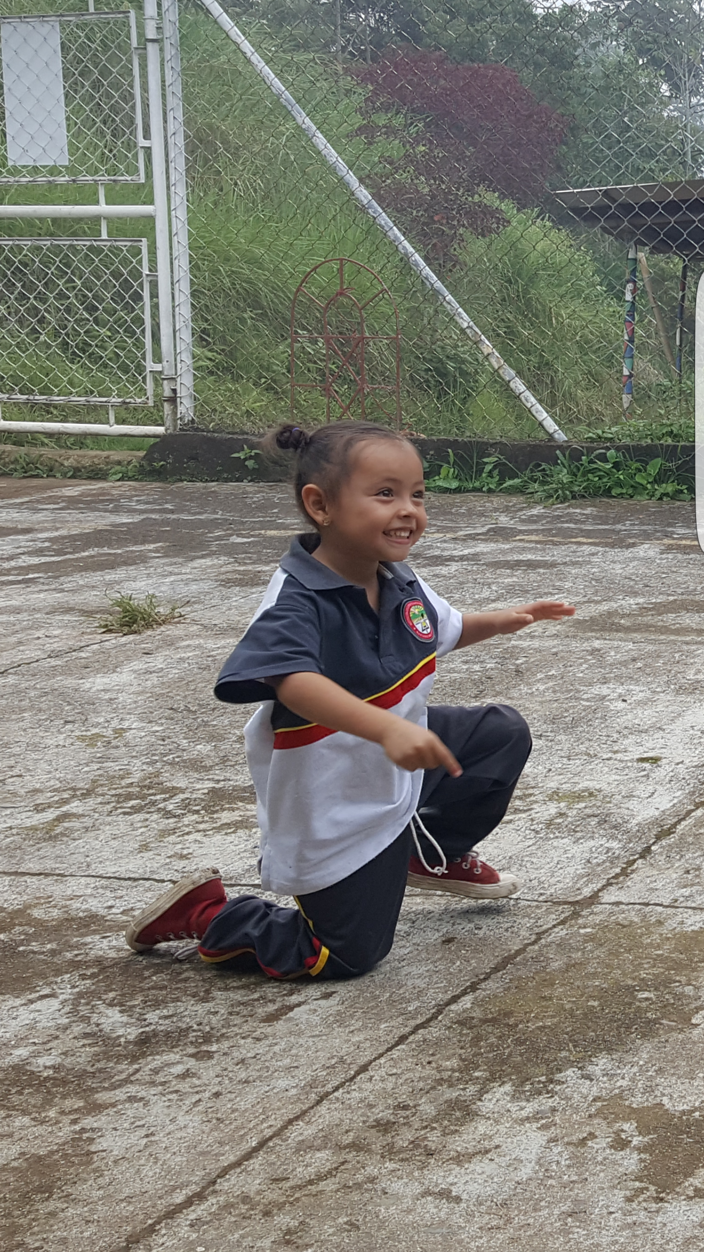 A young girl with dark hair tied in a bun, smiling and kneeling  outdoors, wearing a navy blue, white, and red school uniform and red sneakers.