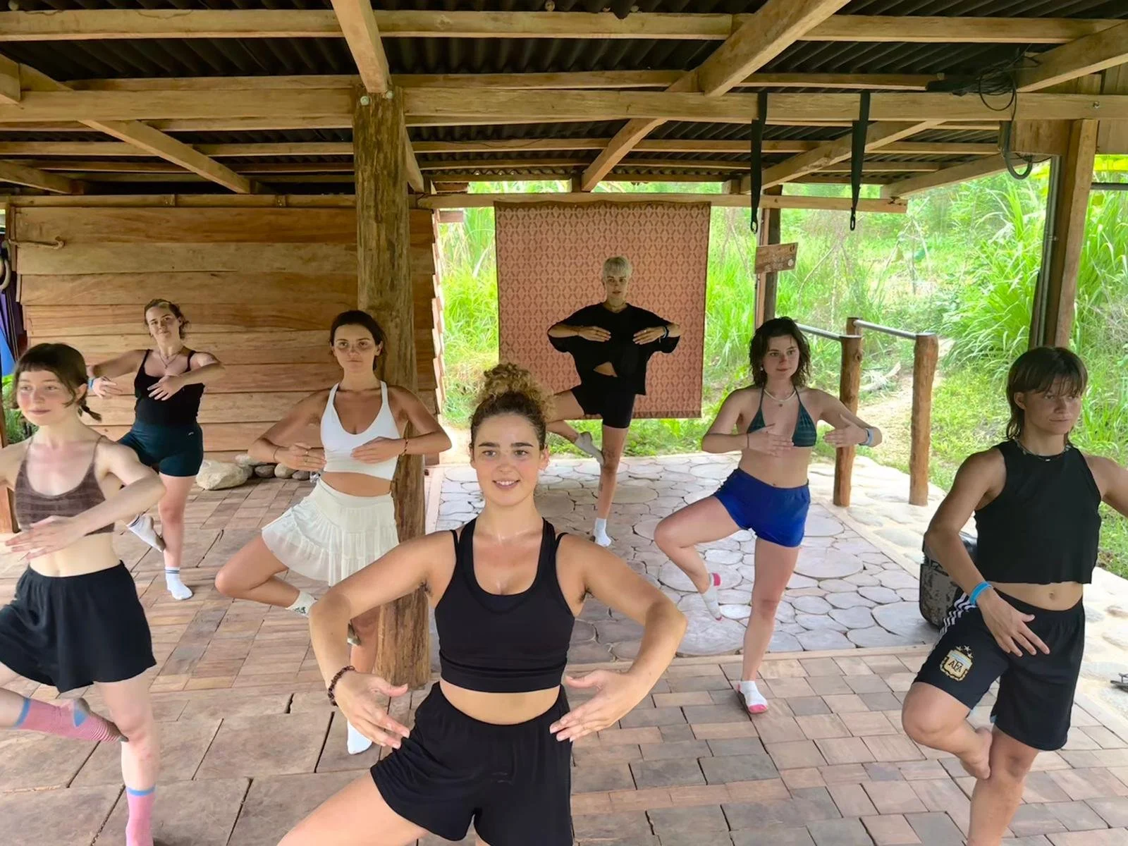 A group of young women in a dance class under a wooden structure surrounded by green foliage, wearing athletic clothing and striking yoga poses.
