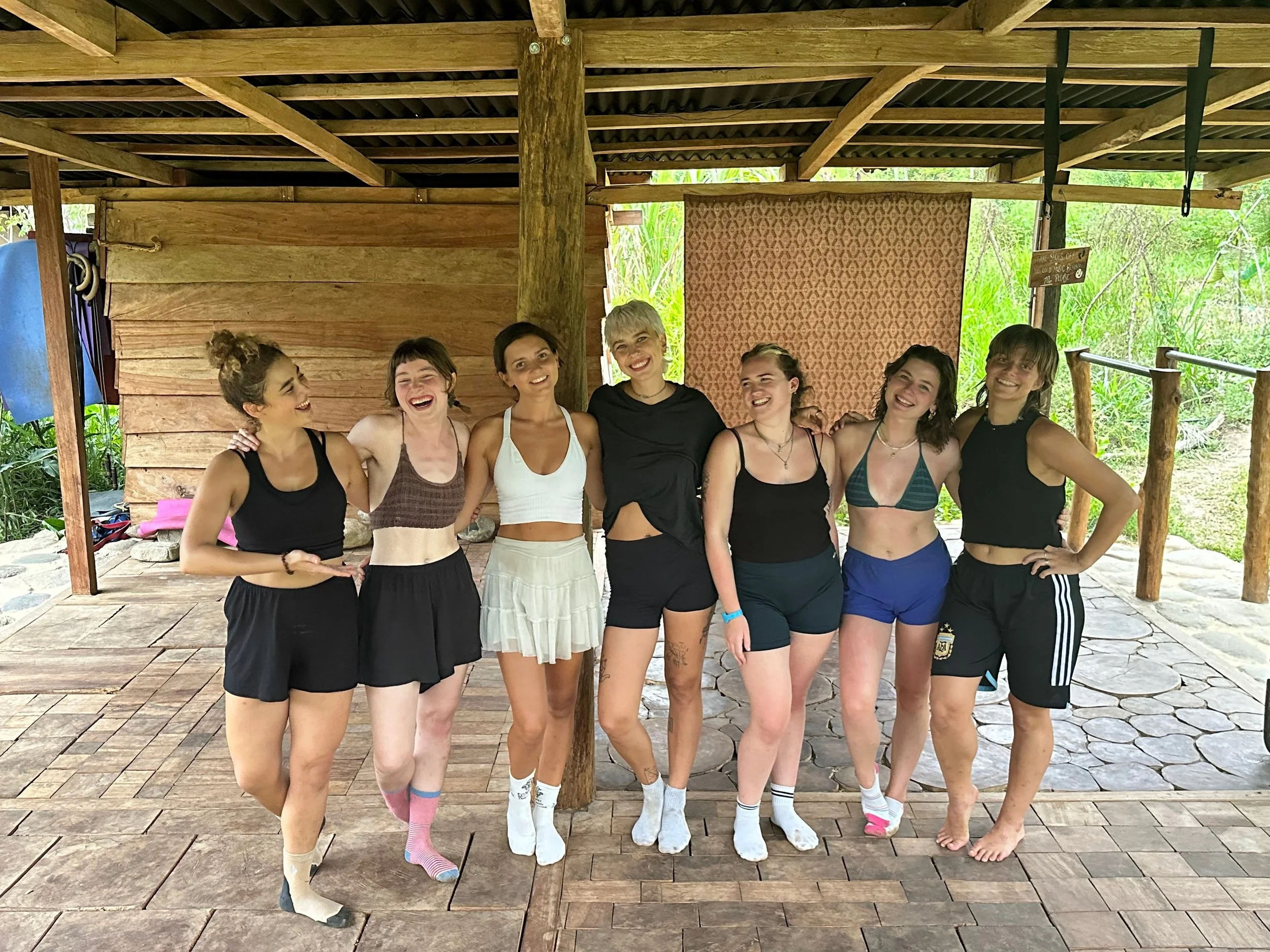 Eight women standing together outdoors under a wooden structure, smiling and hugging each other, dressed in athletic wear, with greenery in the background.