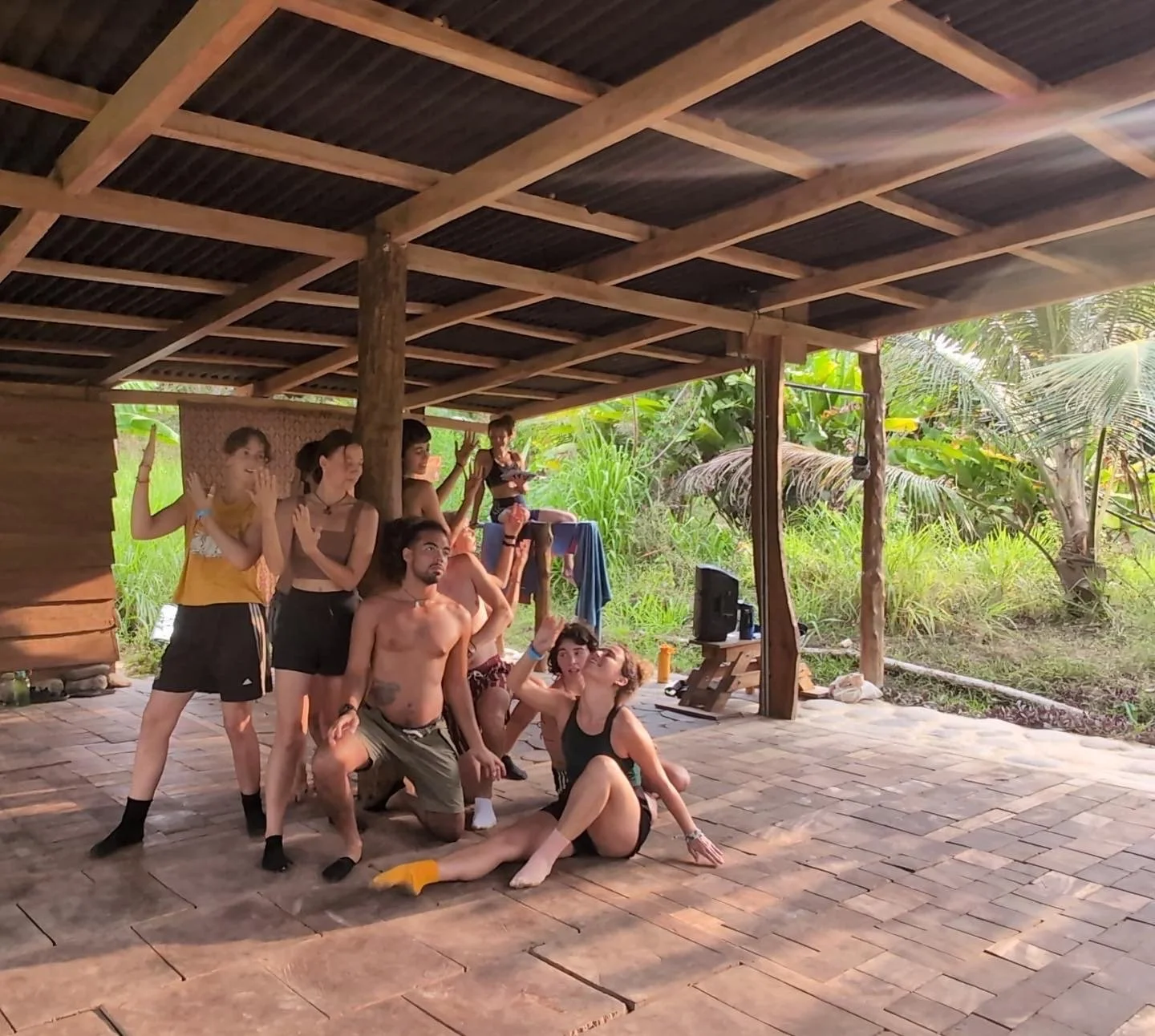 Group of young people posing and enjoying themselves outdoors under a wooden shelter with green foliage in the background.