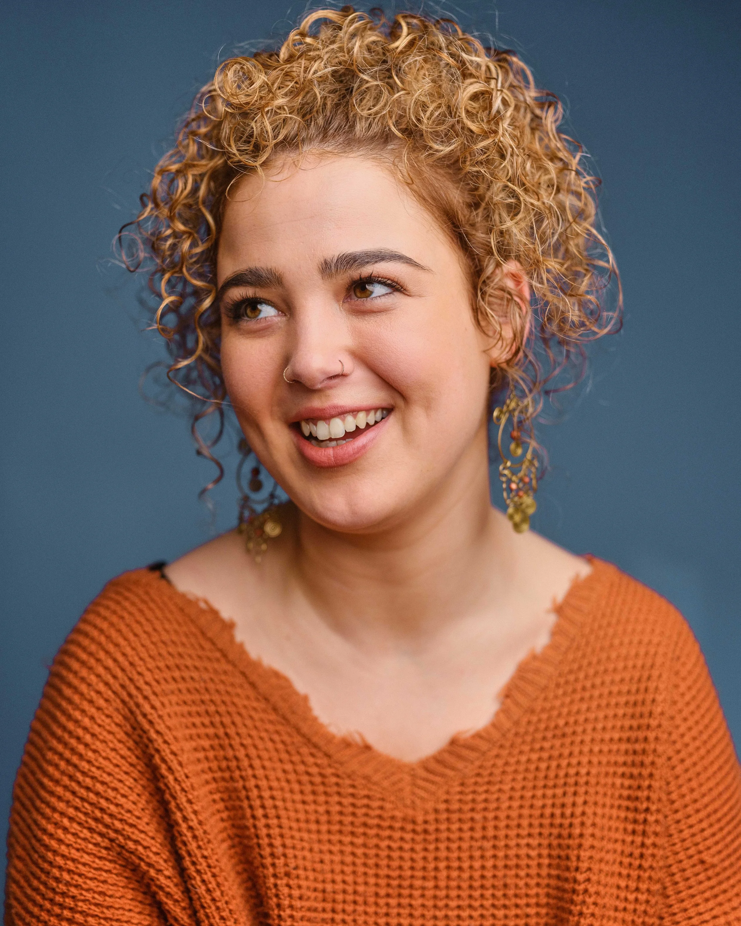 Close-up of a woman with curly blonde hair wearing an orange knitted sweater, smiling and looking to the side against a dark background.