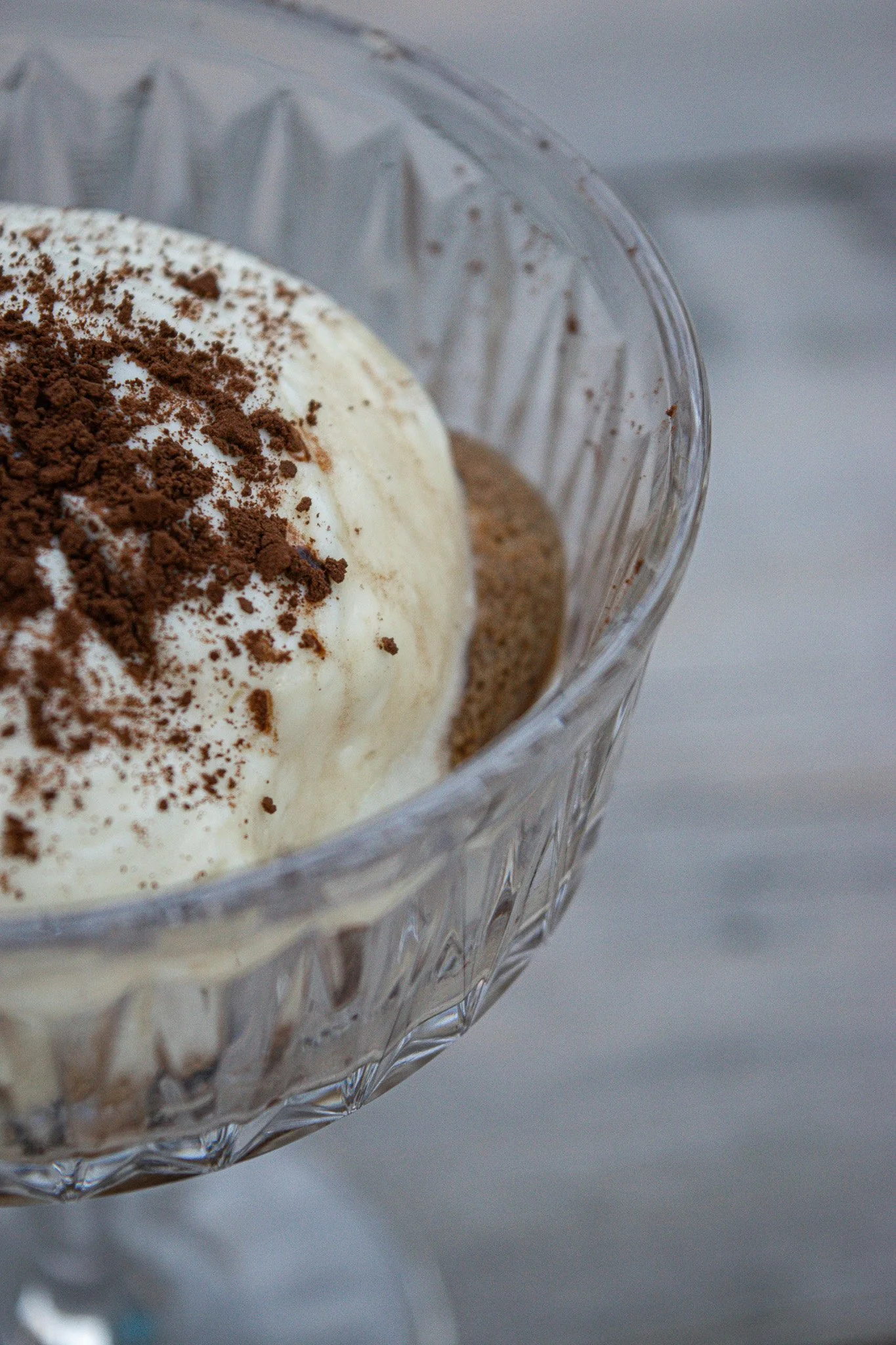 Close-up of a glass dessert dish with whipped cream, cocoa powder, and crushed cookies or chocolate shavings on top.