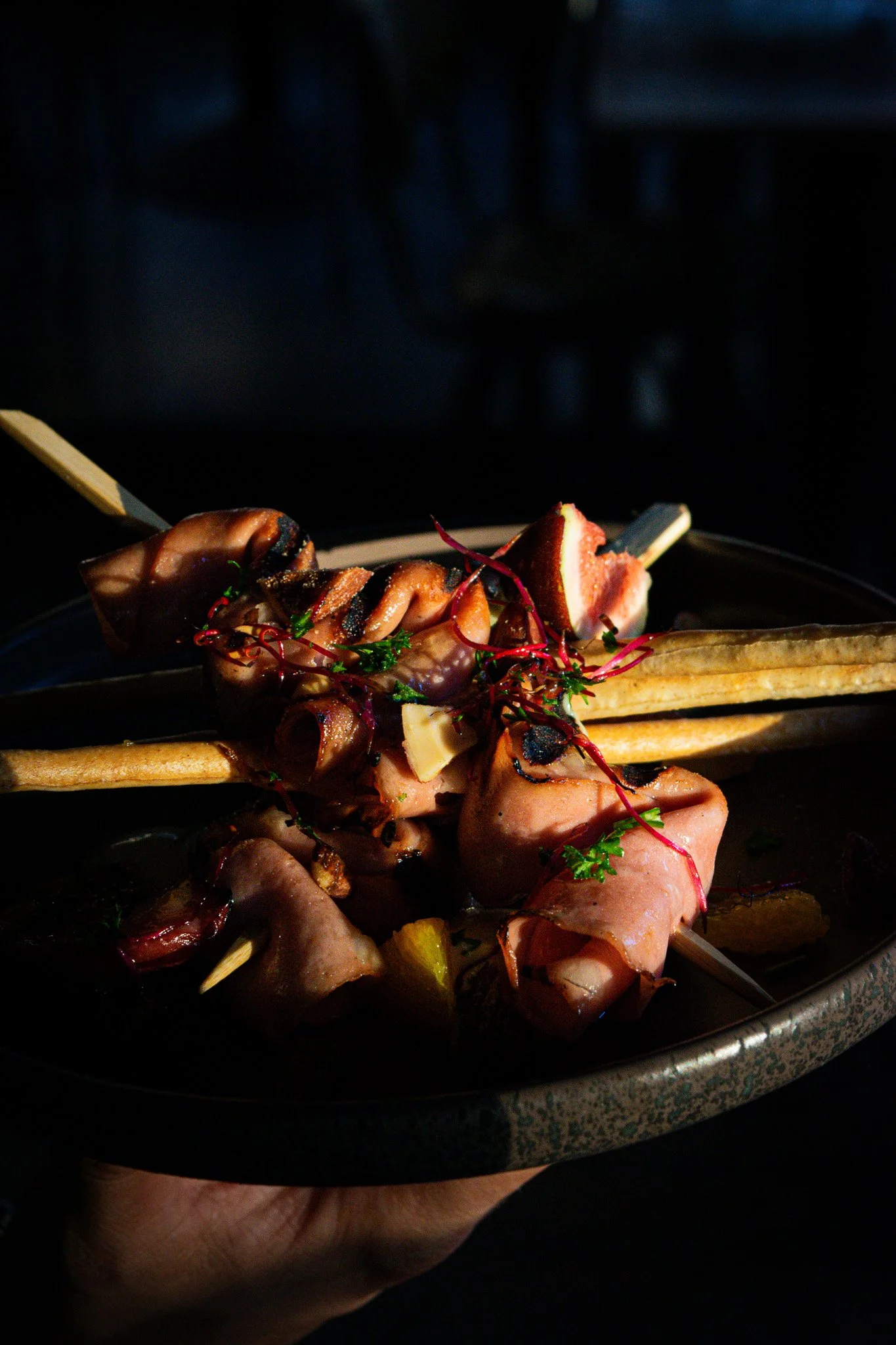 Close-up of a hand holding a black dish with skewered grilled meats, garnished with herbs and vegetables, against a dark background.