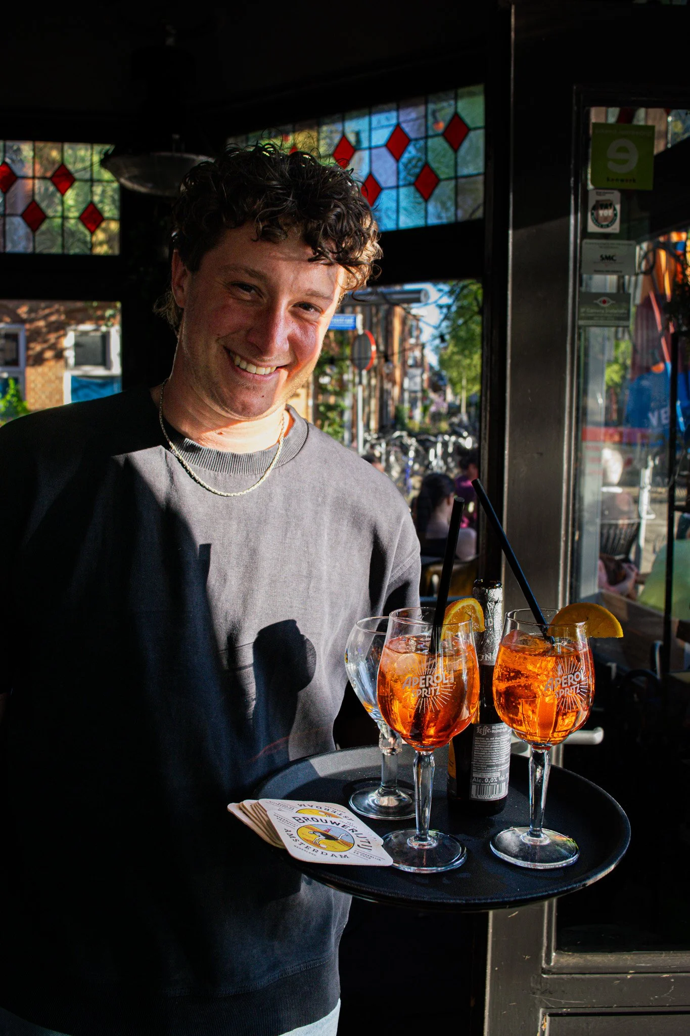 Man die een zwart dienblad met drie Aperol Spritz cocktails vasthoudt in een restaurant of bar, met een menu en een kleurrijke achtergrond met gebrandschilderde ramen.