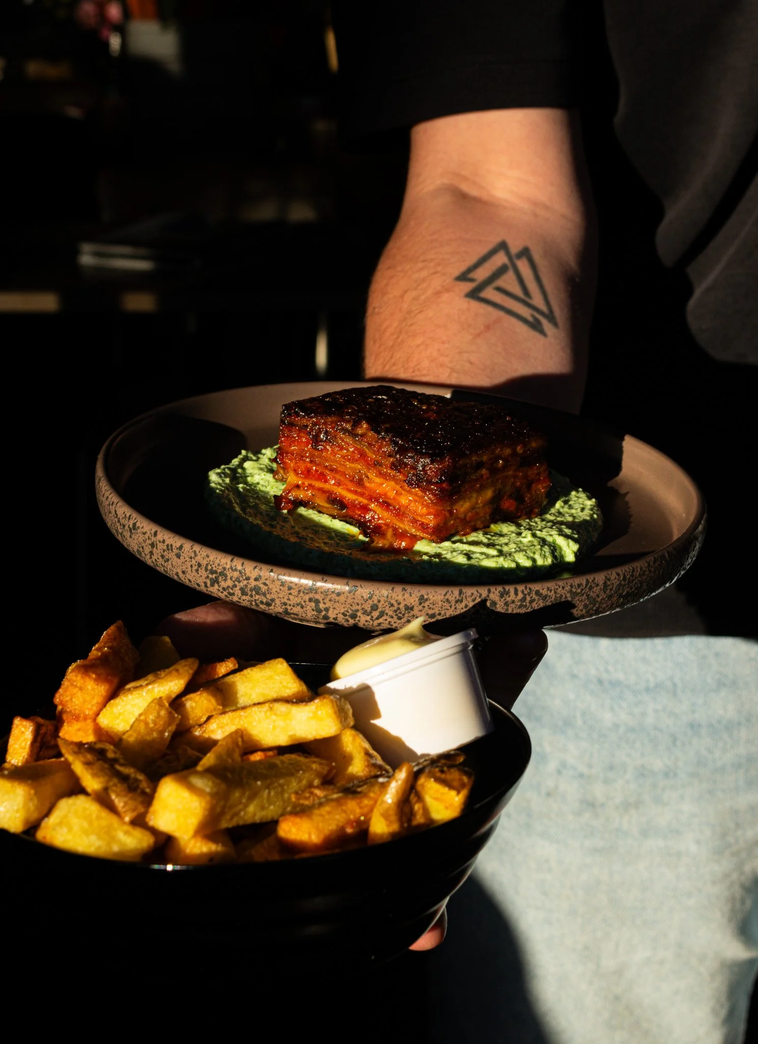 Close-up of a plate with a piece of lasagna on top of a green sauce, and a side of French fries with mayonnaise in a small cup. A person's arm with a tattoo is in the background.