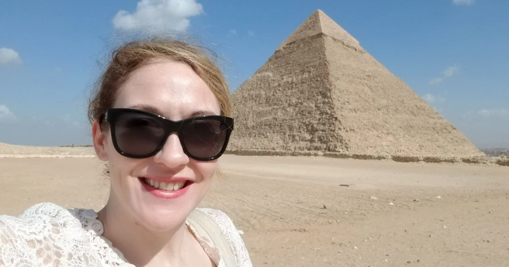 A woman with sunglasses and a white lace top smiling for a selfie in front of the Great Pyramid of Giza under a blue sky with scattered clouds.