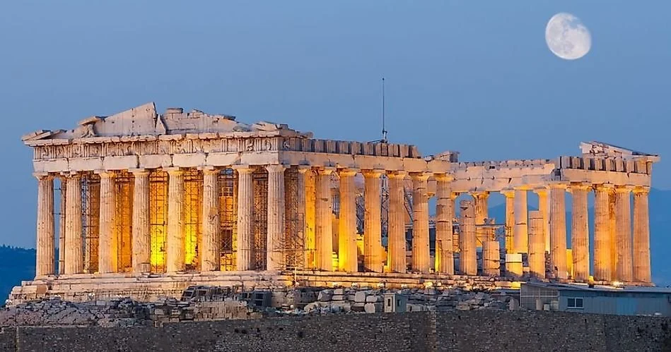 The Parthenon, an ancient Greek temple, illuminated at night with a full moon in the sky above.