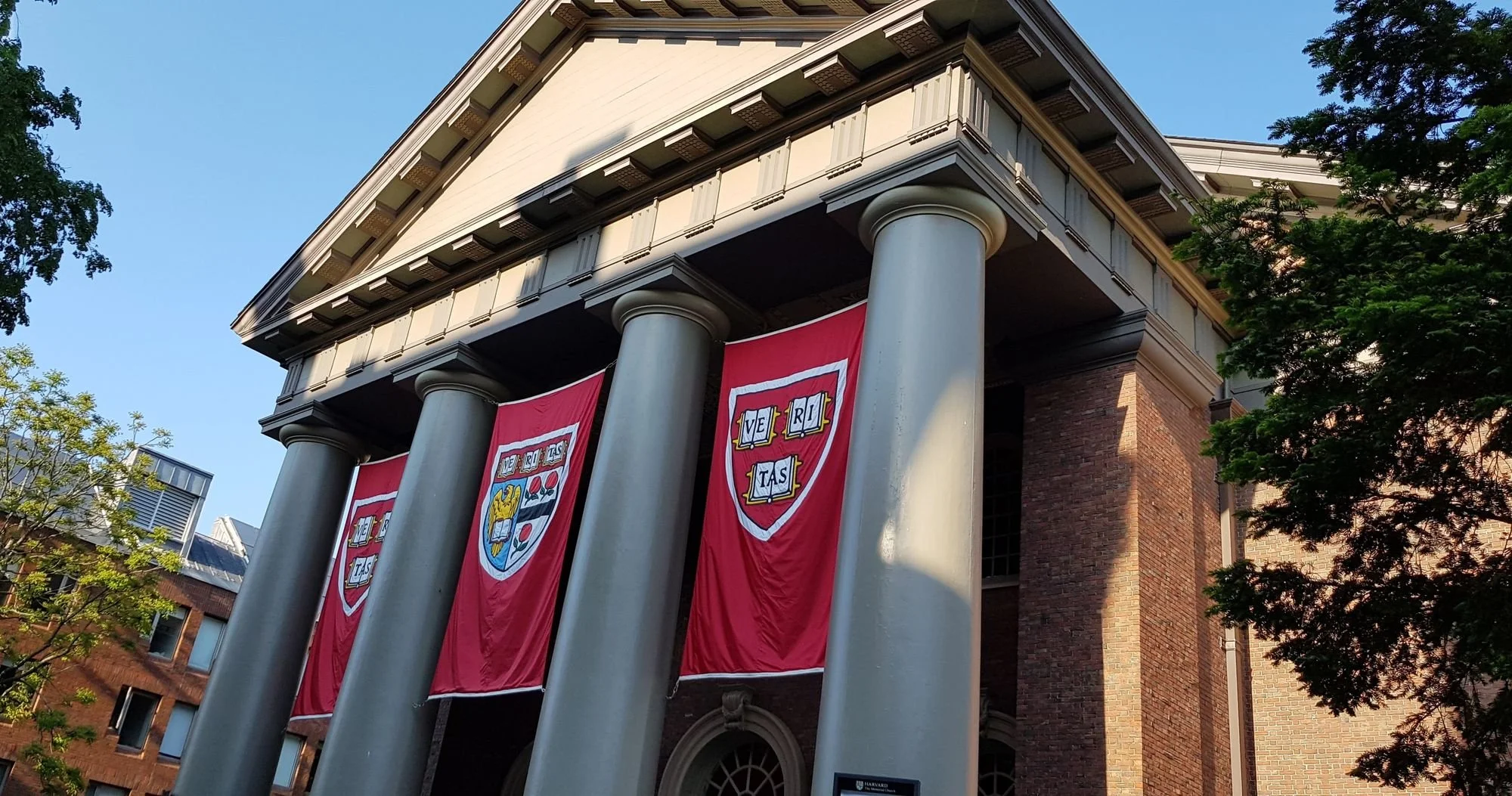 Main entrance to Widener Library at Harvard University and Harvard banners hanging from them, trees on the sides, and a clear daytime sky.
