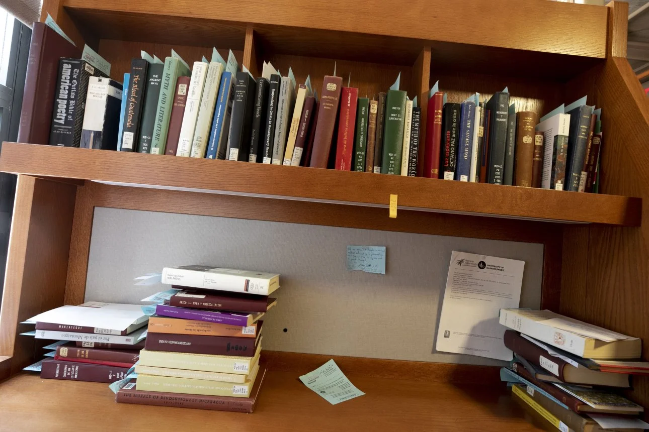 A wooden bookshelf filled with books on the upper shelves, and a cluttered workspace with stacked books, papers, and sticky notes on the lower desk area.