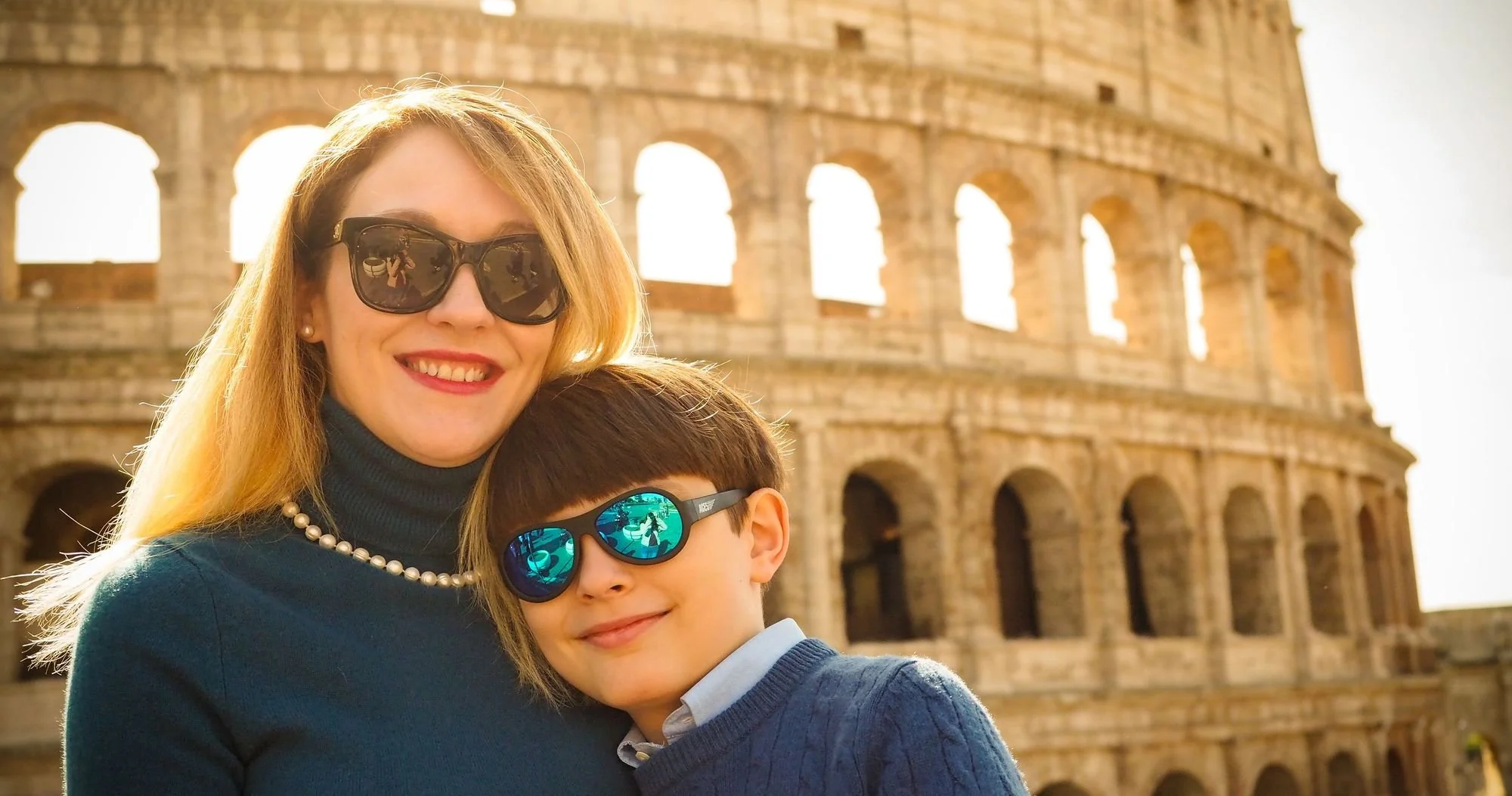 A woman and a young boy, both wearing sunglasses, smiling in front of the Colosseum in Rome, Italy.