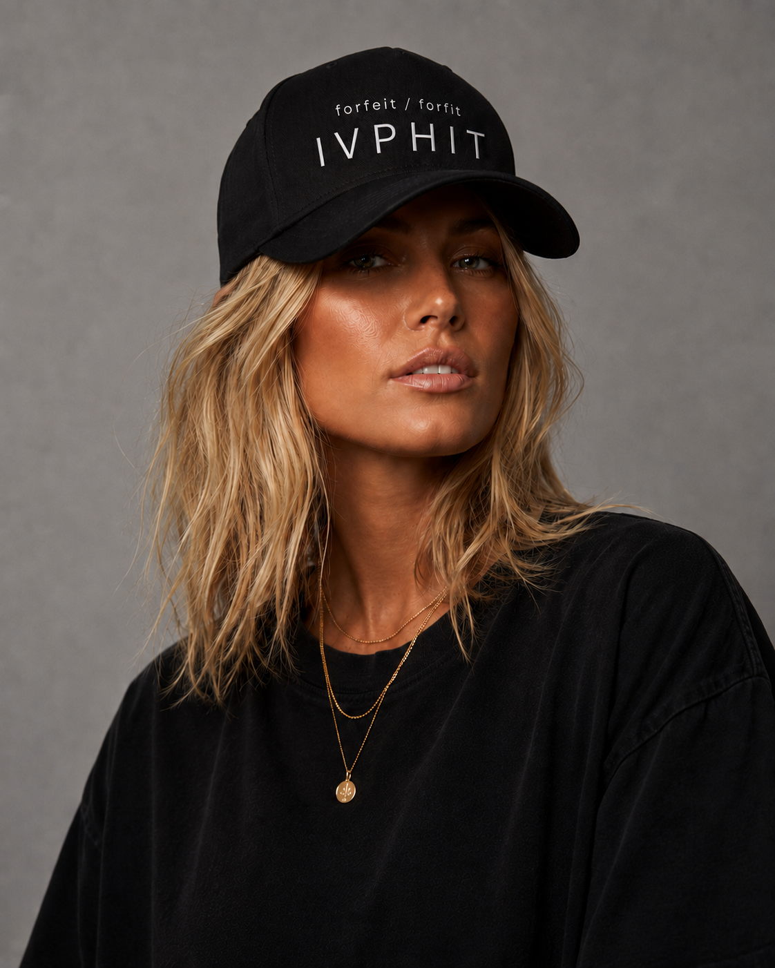 A woman wearing a black baseball cap with white text, a black shirt, and gold layered necklaces. She has blonde, wavy hair and a neutral expression, posing against a gray background.
