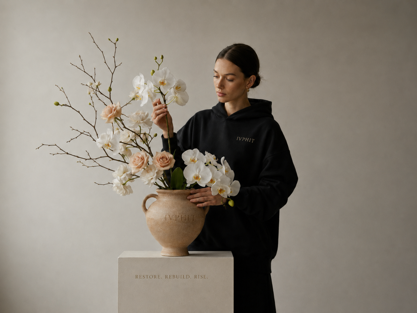 A woman in a black hoodie arranging white orchids and pale pink roses in a large beige ceramic vase with the word 'IVPHIT' embossed on it, on a white pedestal with the words 'Restore. Rebuild. Rise.' written on it.