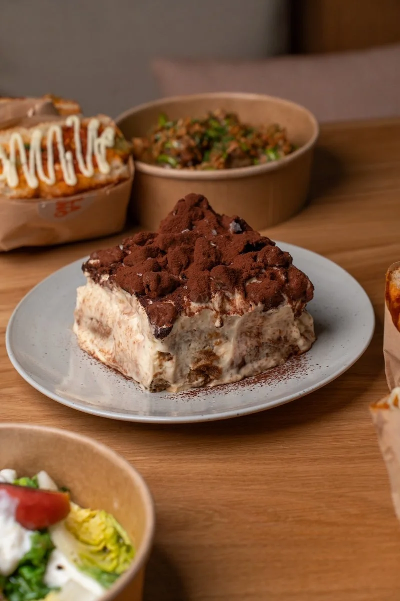 Ovomaltine tiramisu from Alla Mano with focaccia sandwiches and loaded patatine on a wooden table.